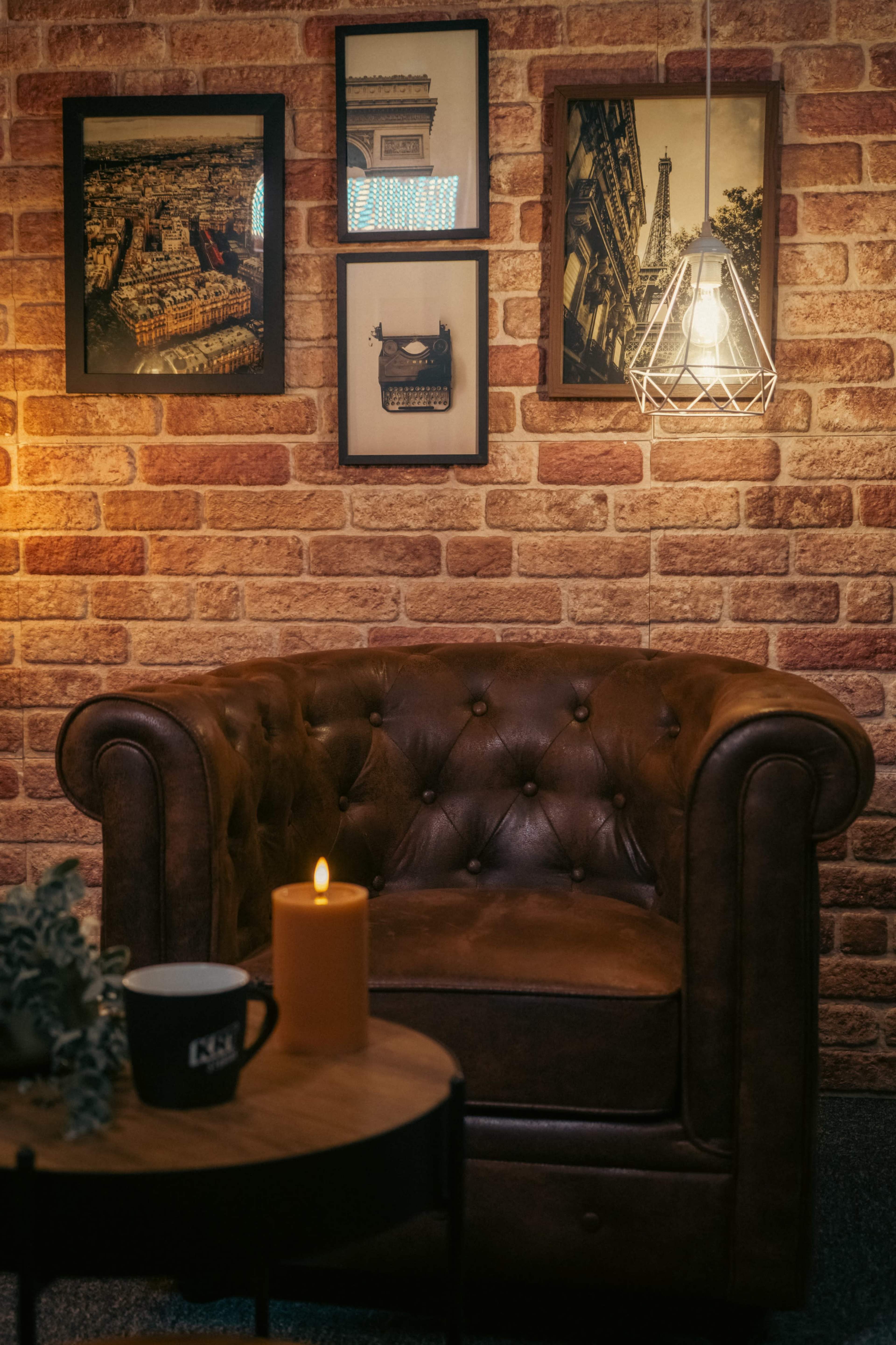 The image depicts a cozy interior setting with a brown leather chair, a small round table, a lit candle, and framed wall art against a brick backdrop.