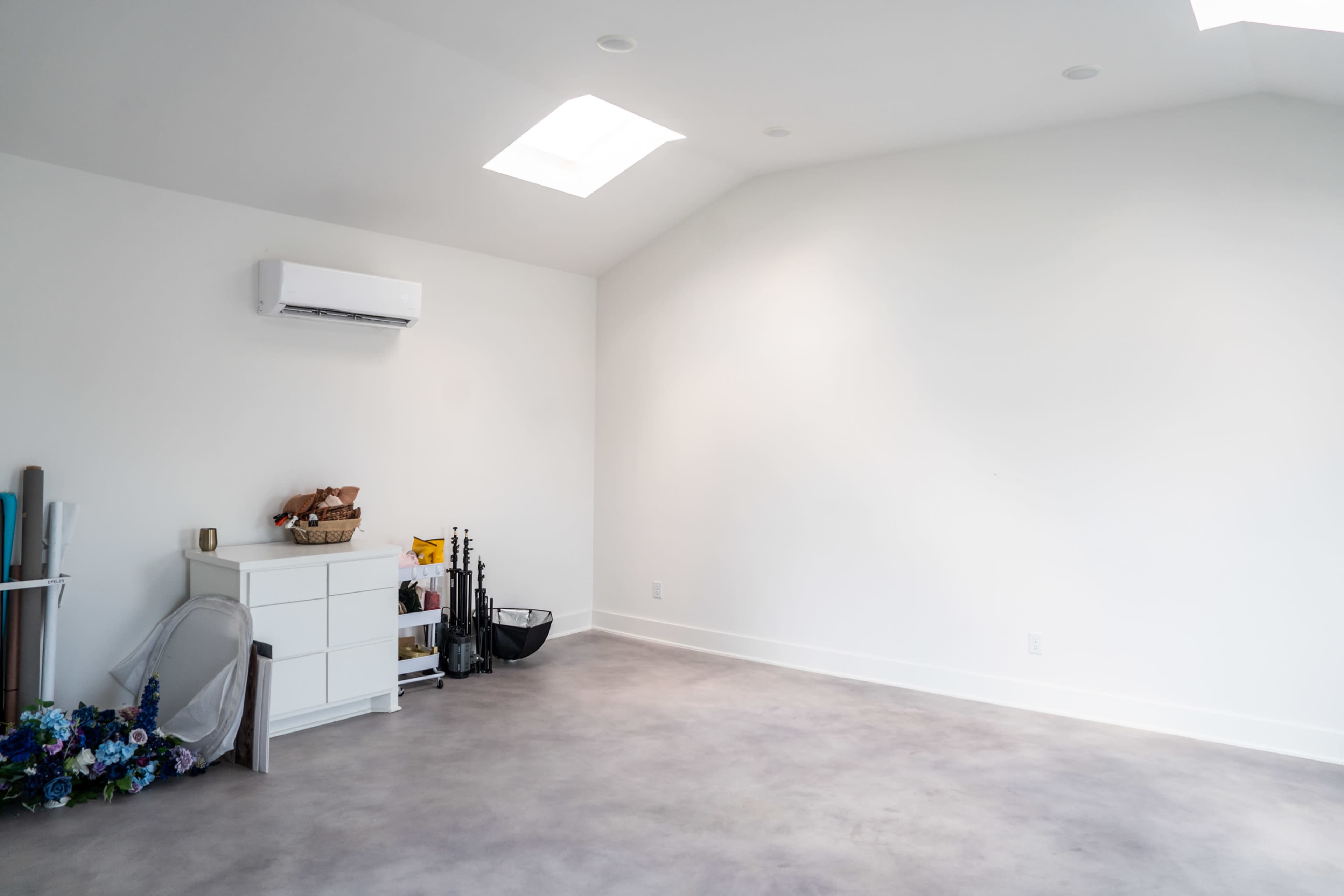 The image shows a spacious, empty room with a high ceiling, a white air conditioning unit, a white dresser, and various items neatly arranged in the corner.