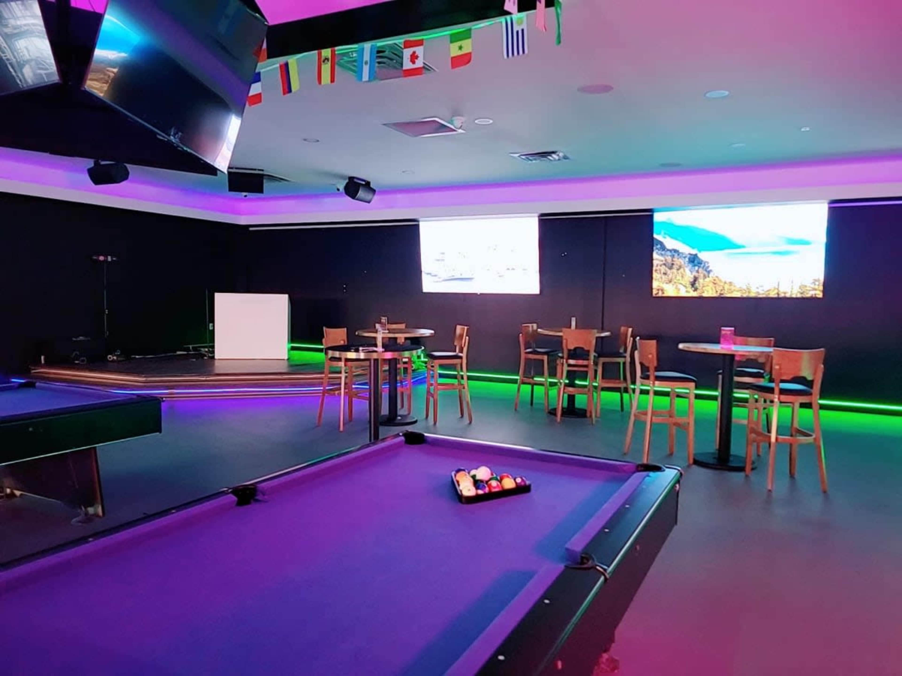 A modern game room features a pool table, high-top tables, and wall-mounted screens, illuminated by colorful lighting and decorated with flags from various countries.
