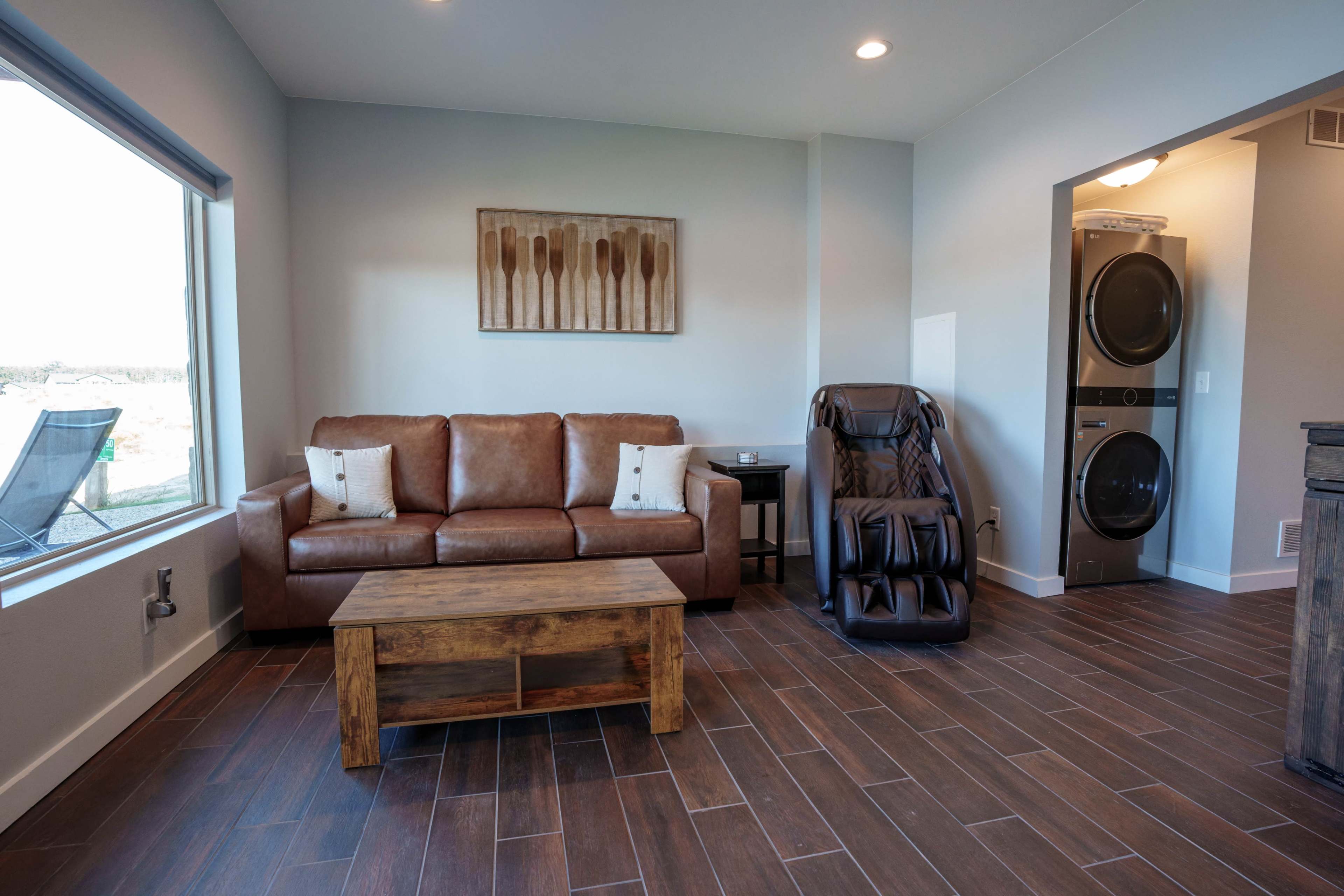 The room features a brown leather sofa, a wooden coffee table, a massage chair, and a stacked washer and dryer in a well-lit space with large windows.