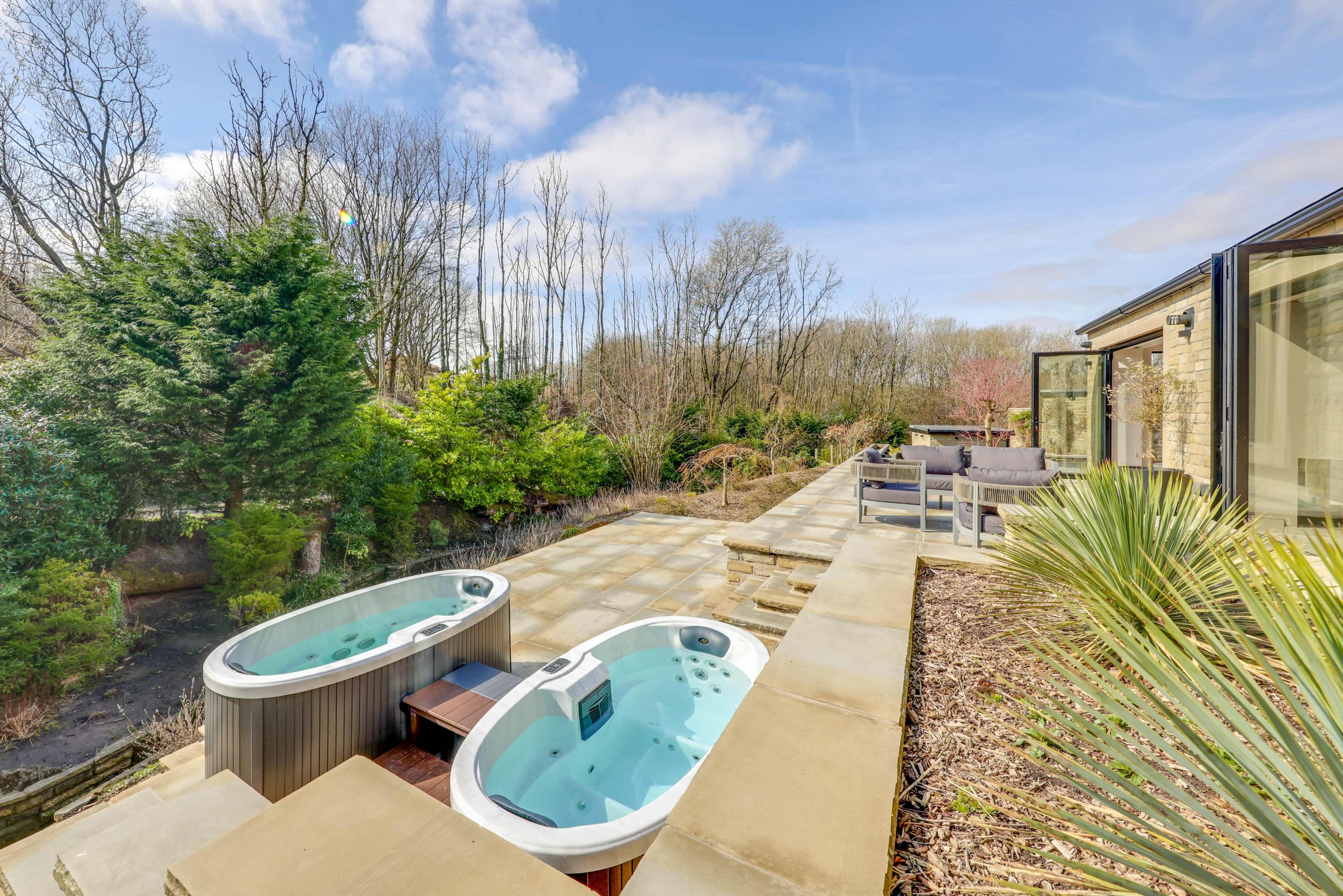 The image shows two hot tubs situated on a stone patio surrounded by greenery and trees.