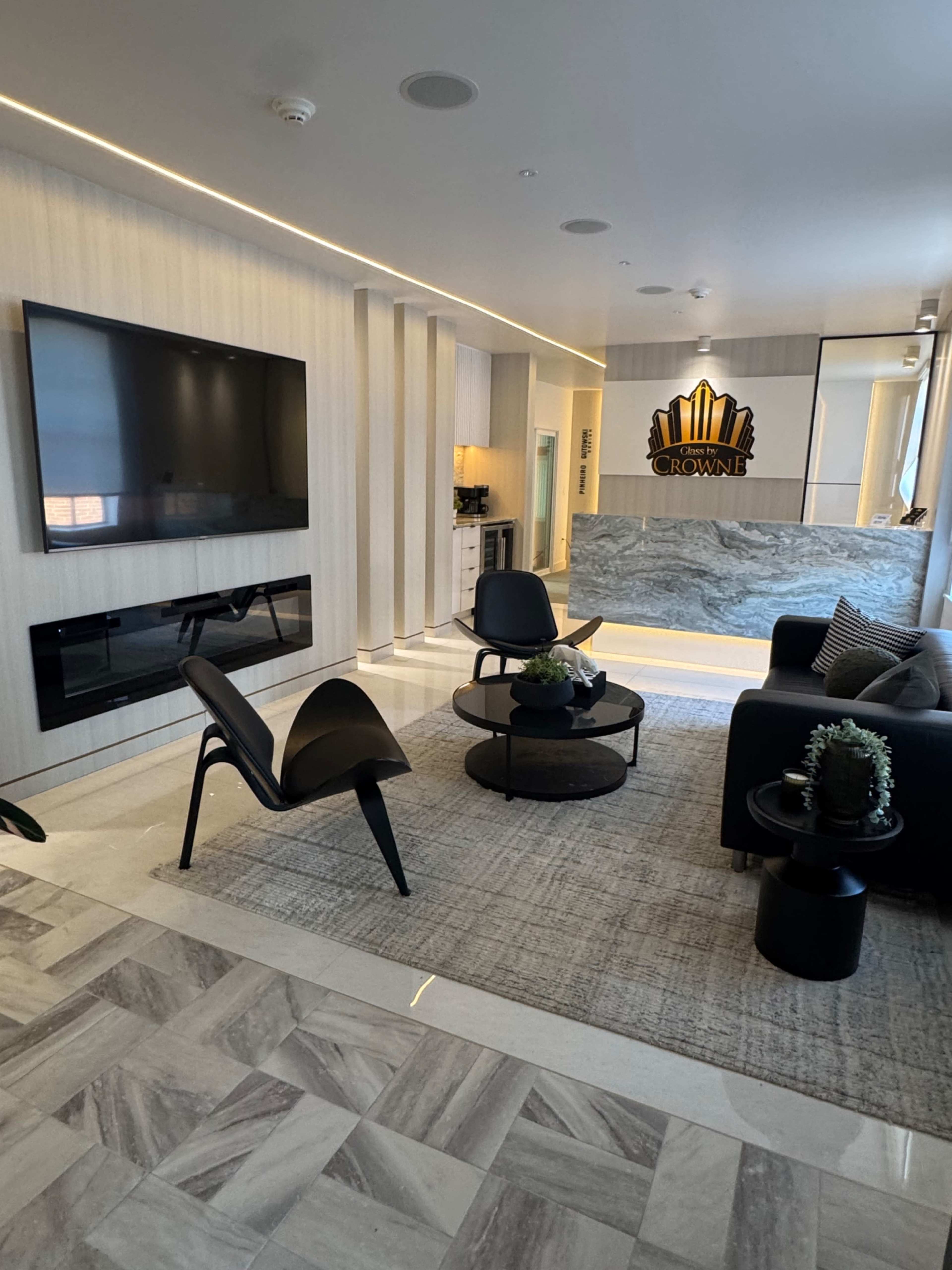 The image shows a modern living room with a large flat-screen TV mounted on a wall, a black coffee table, a dark sofa, and two chairs arranged on a light gray rug over tiled flooring.