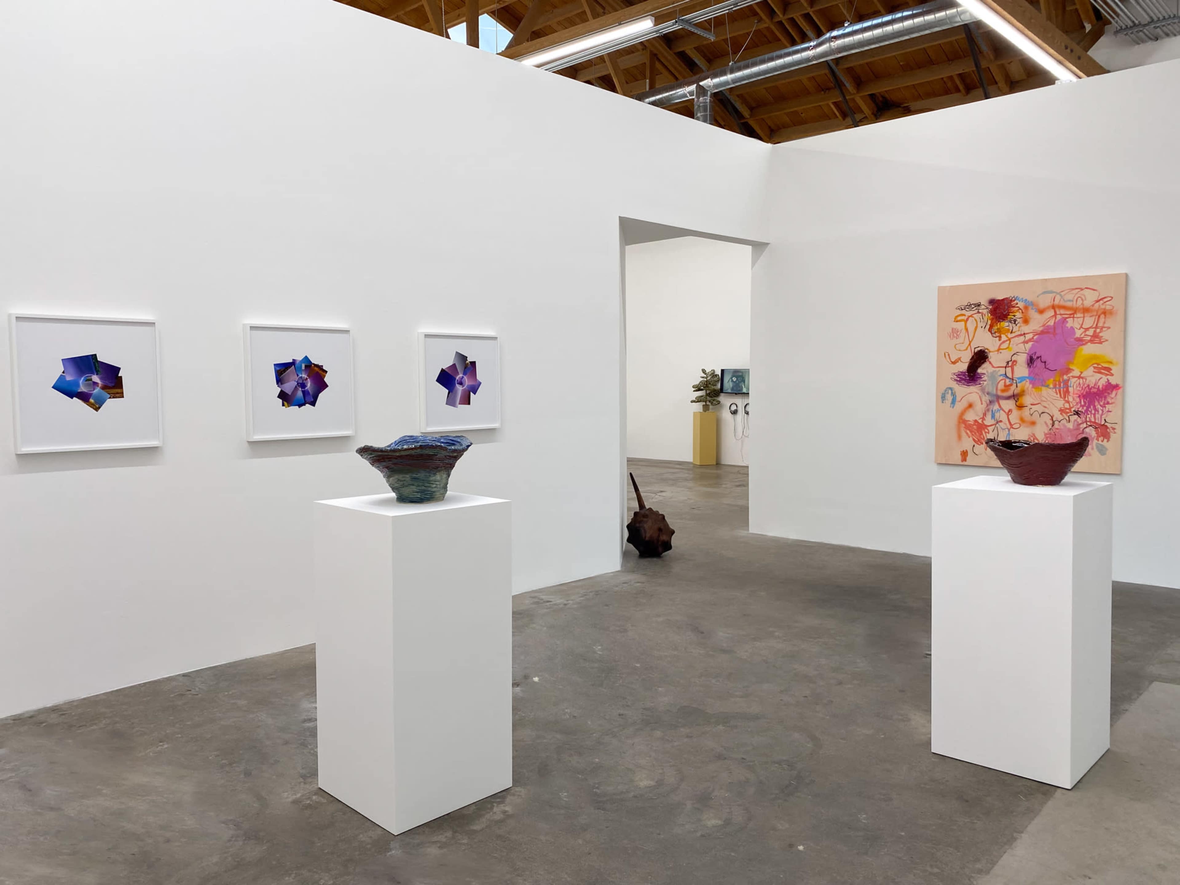 An art gallery featuring four framed artworks on a white wall, two decorative bowls on pedestals, and a colorful painting on an adjacent wall.