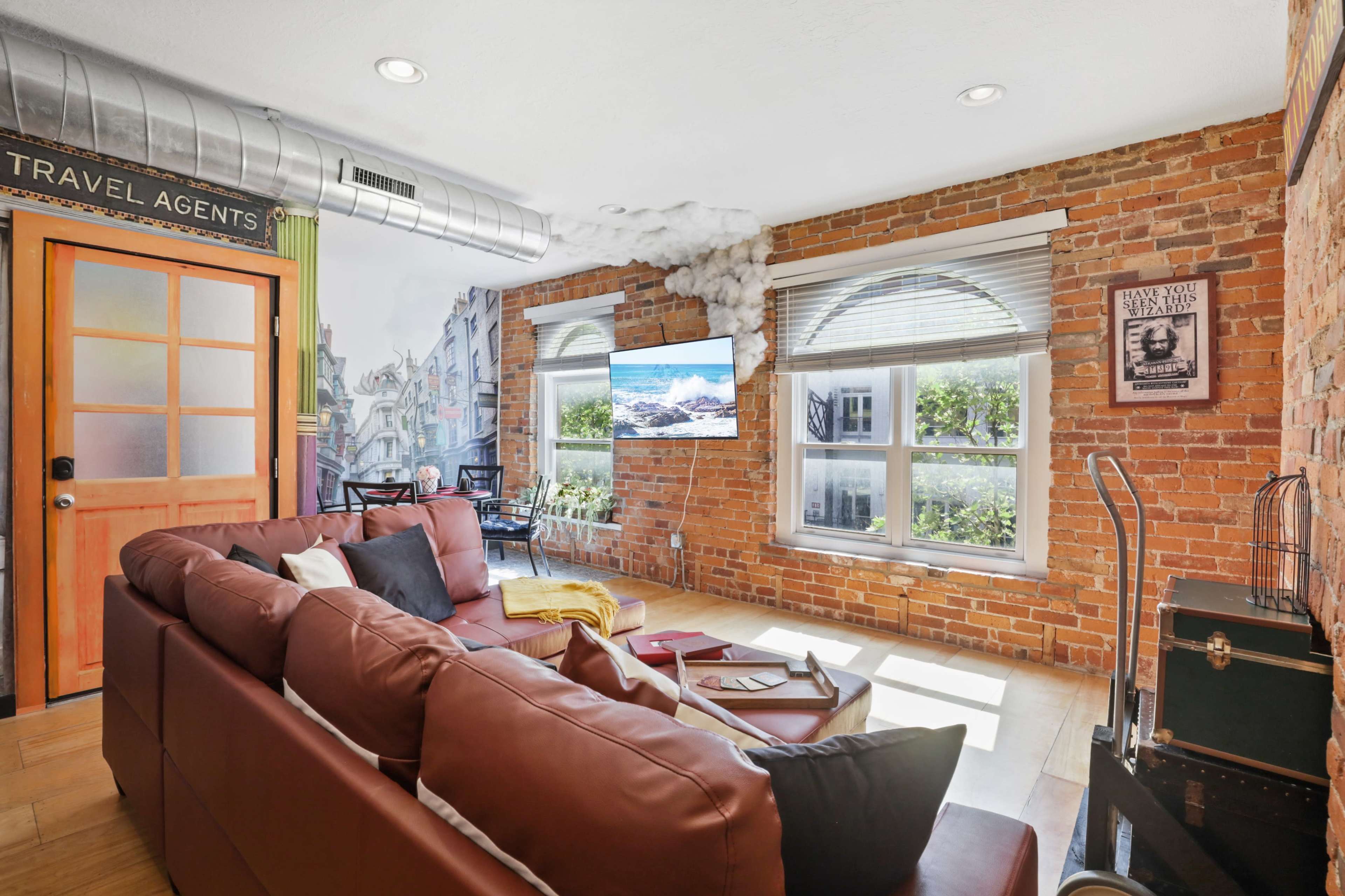 A cozy living room features a brown leather couch, large windows with city views, and exposed brick walls.