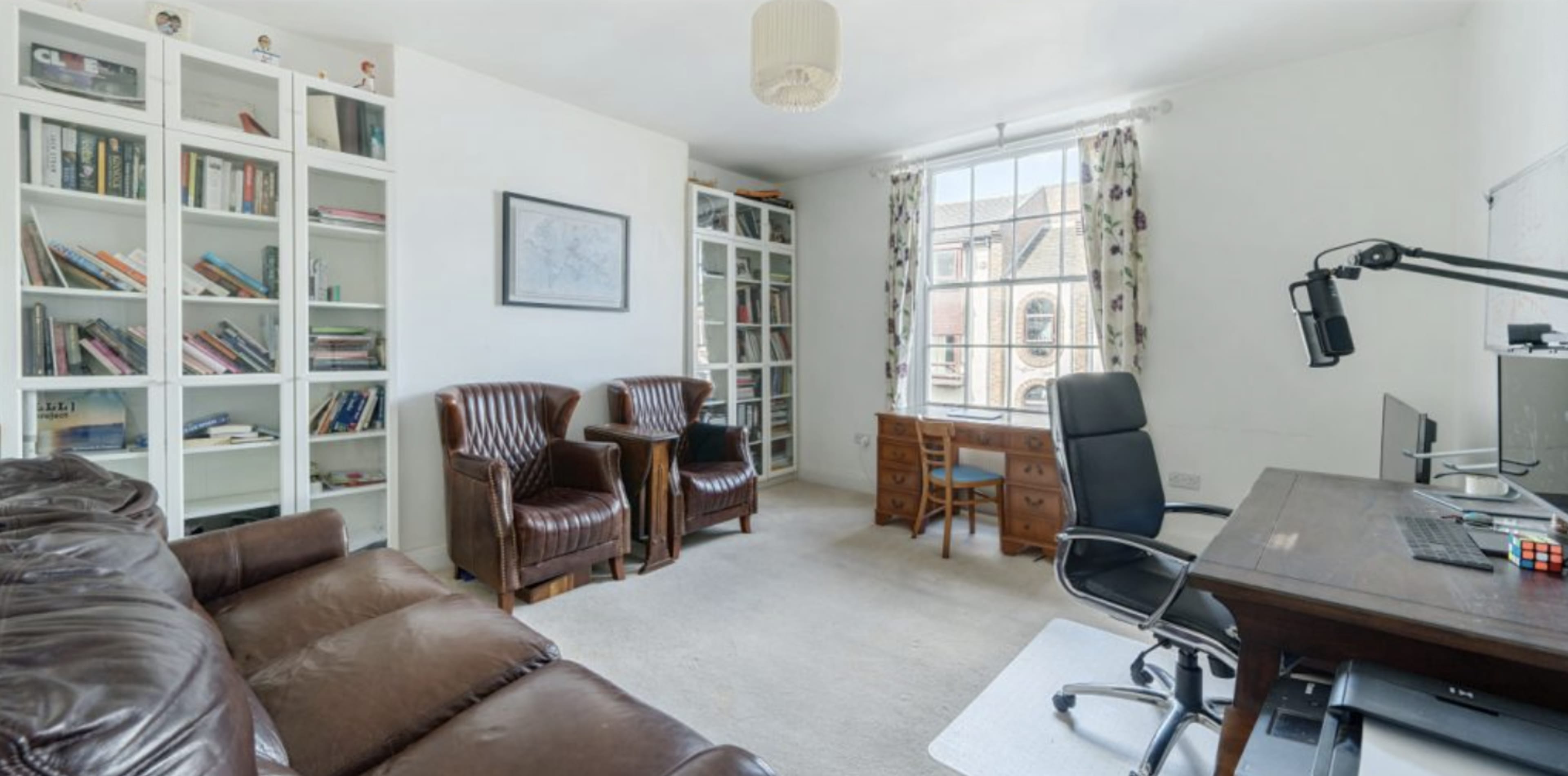 The image shows a well-lit home office with a desk, a computer, two leather armchairs, and bookshelves filled with books.