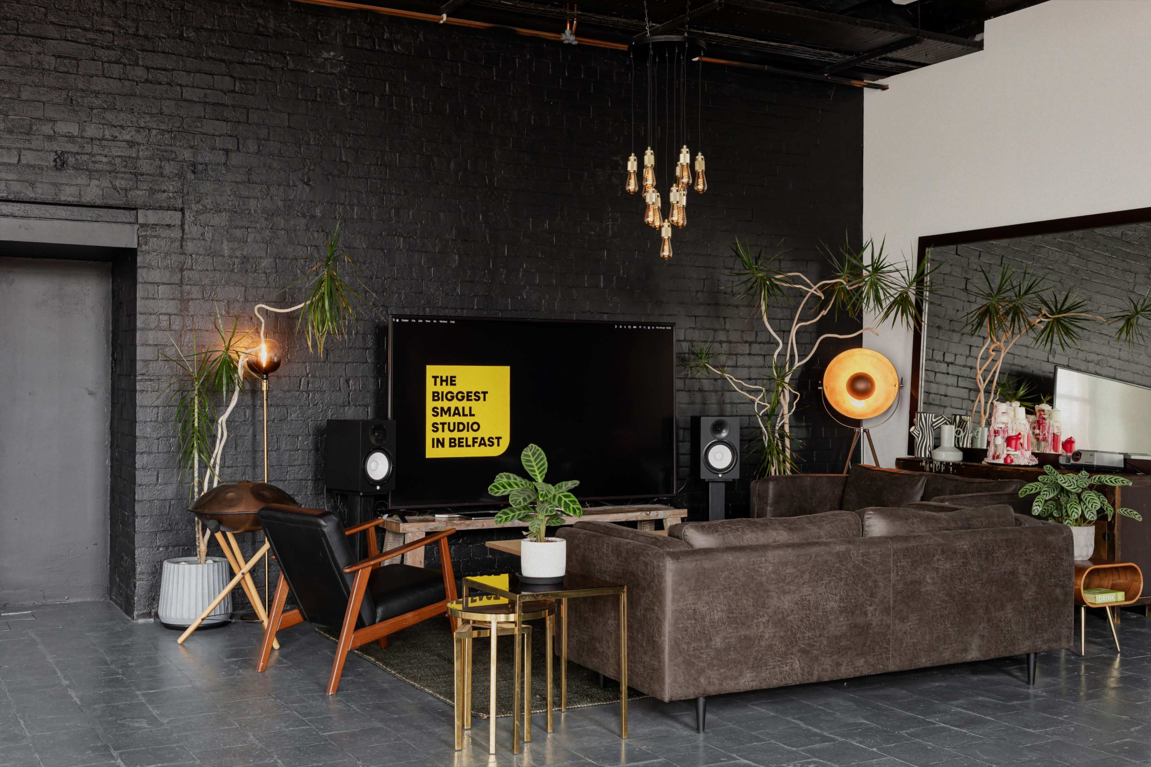 The image shows a modern living room with a large black television displaying text, a brown sofa, plants, and stylish lighting fixtures against a dark brick wall.