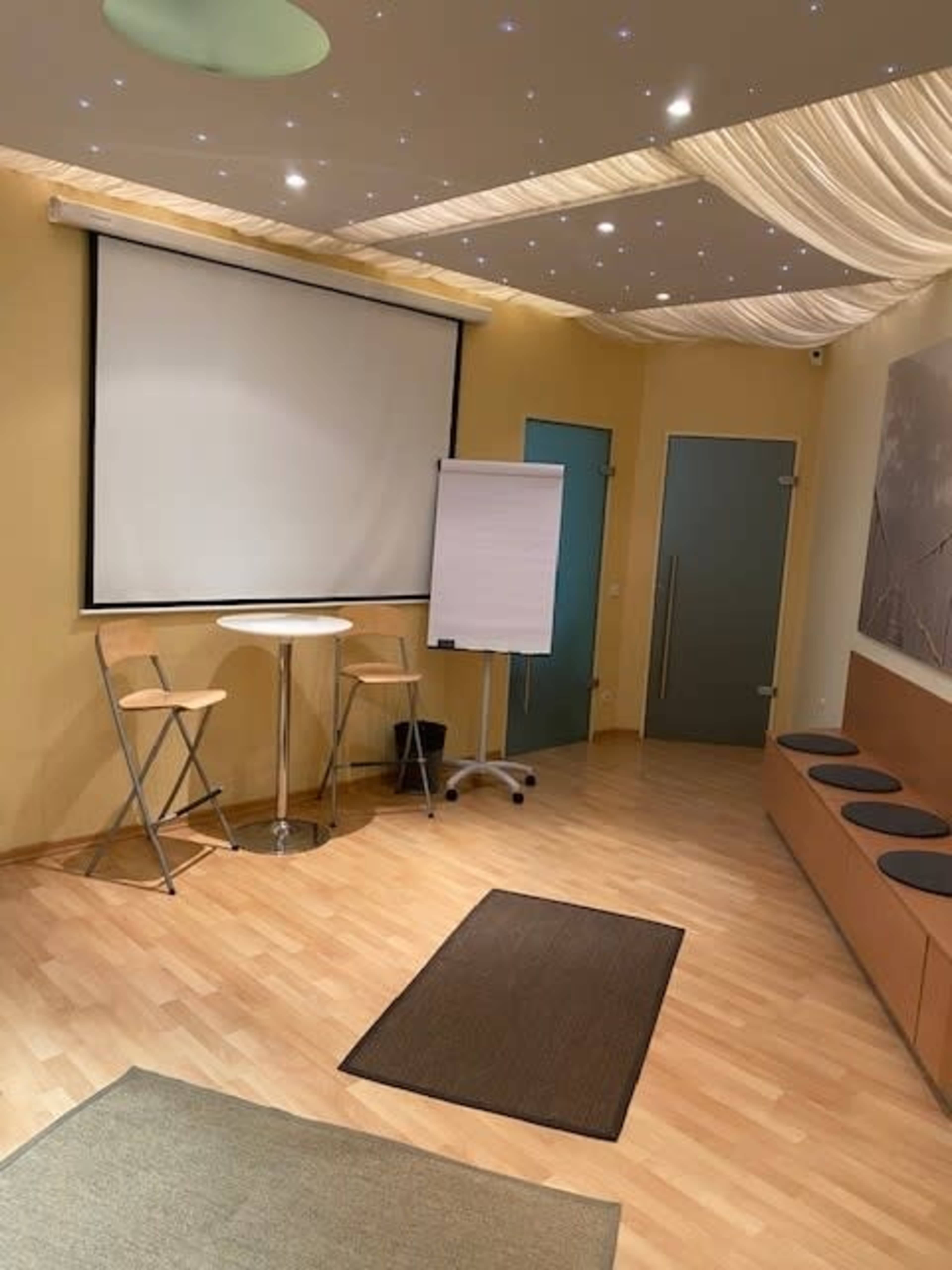 A meeting room featuring a large screen, a flip chart, two high stools, and a bench along the wall, with soft lighting and wooden flooring.