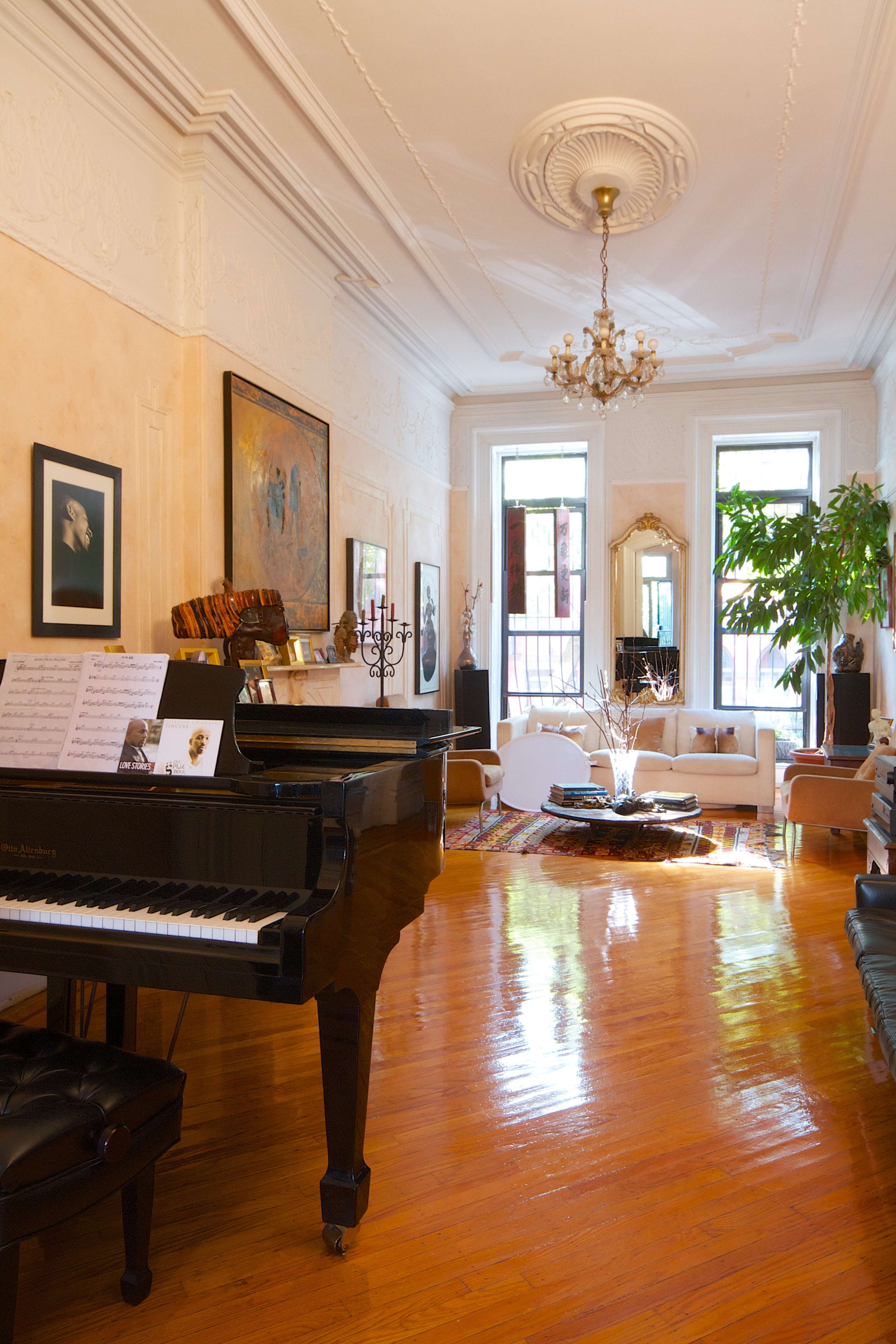 The image shows a spacious room with a grand piano on one side, a chandelier hanging from the ceiling, and a large window allowing sunlight to illuminate a seating area with a plant.