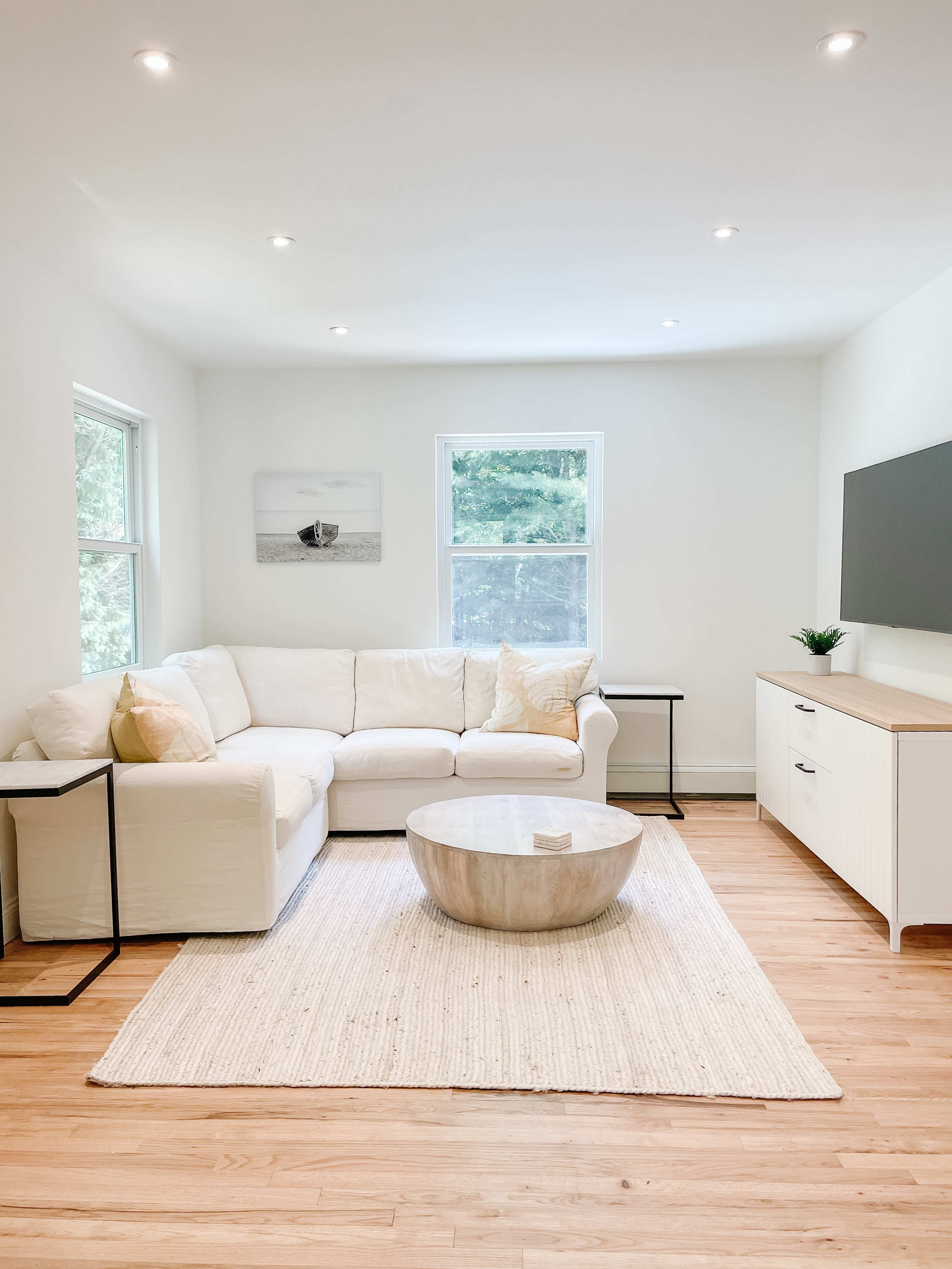 A bright, minimalist living room with a white sectional sofa, a round coffee table, and a TV mounted on the wall.