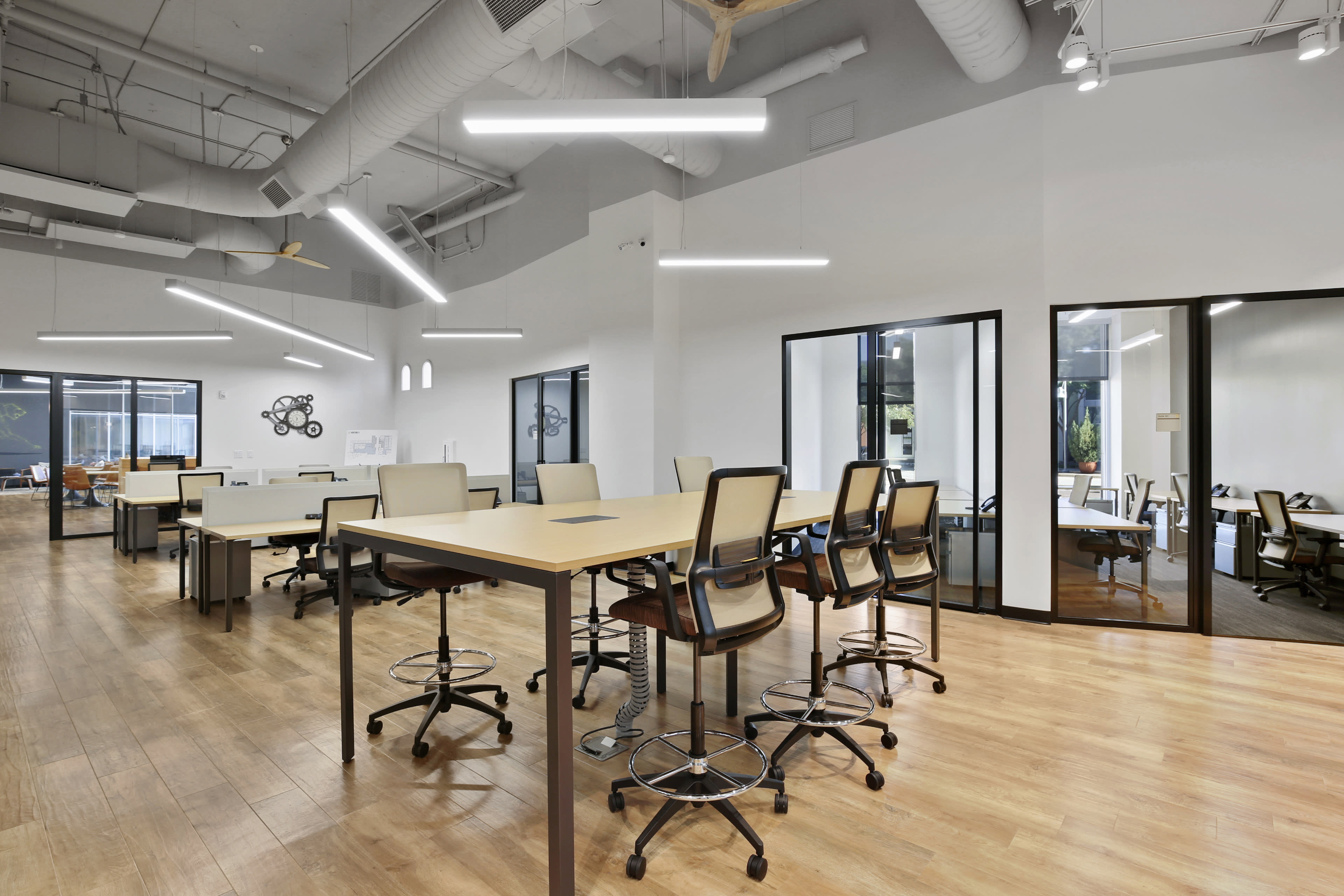 The image shows a modern office space with multiple workstations, featuring desks and ergonomic chairs, along with glass-enclosed meeting rooms in the background.