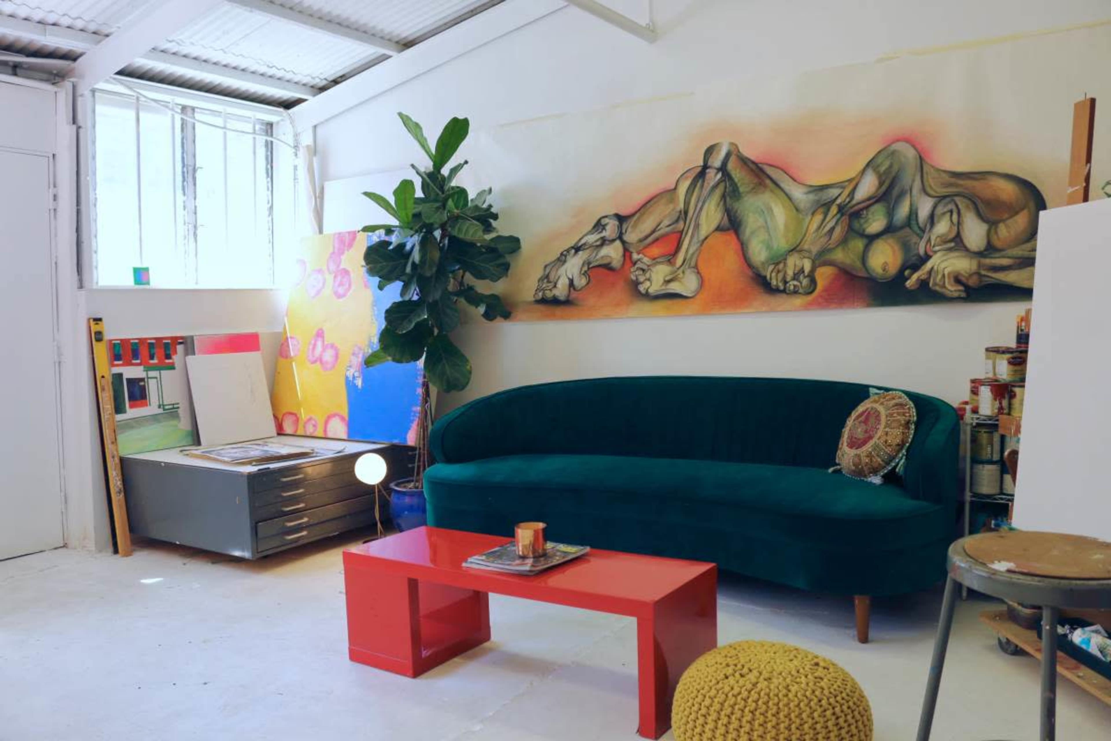 A cozy art studio featuring a green sofa, a red coffee table, and a large colorful mural on the wall, surrounded by art supplies and canvases.