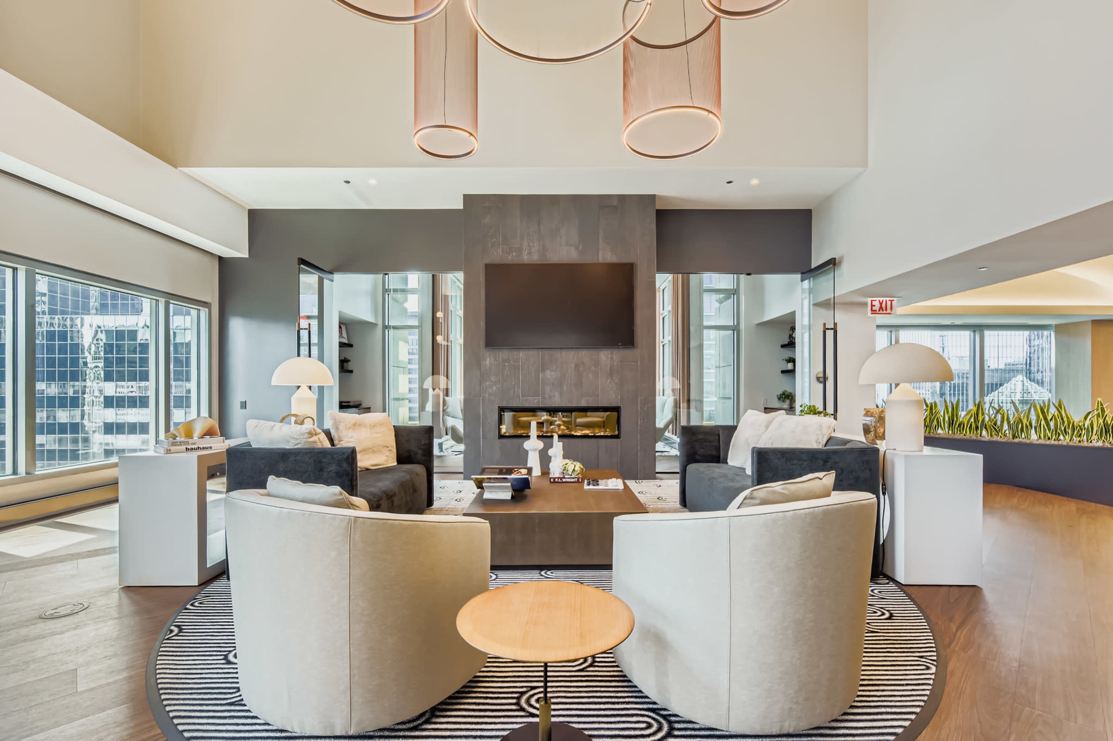 The image shows a modern lounge area with two plush chairs facing a central coffee table, a fireplace, and large windows revealing a cityscape outside.
