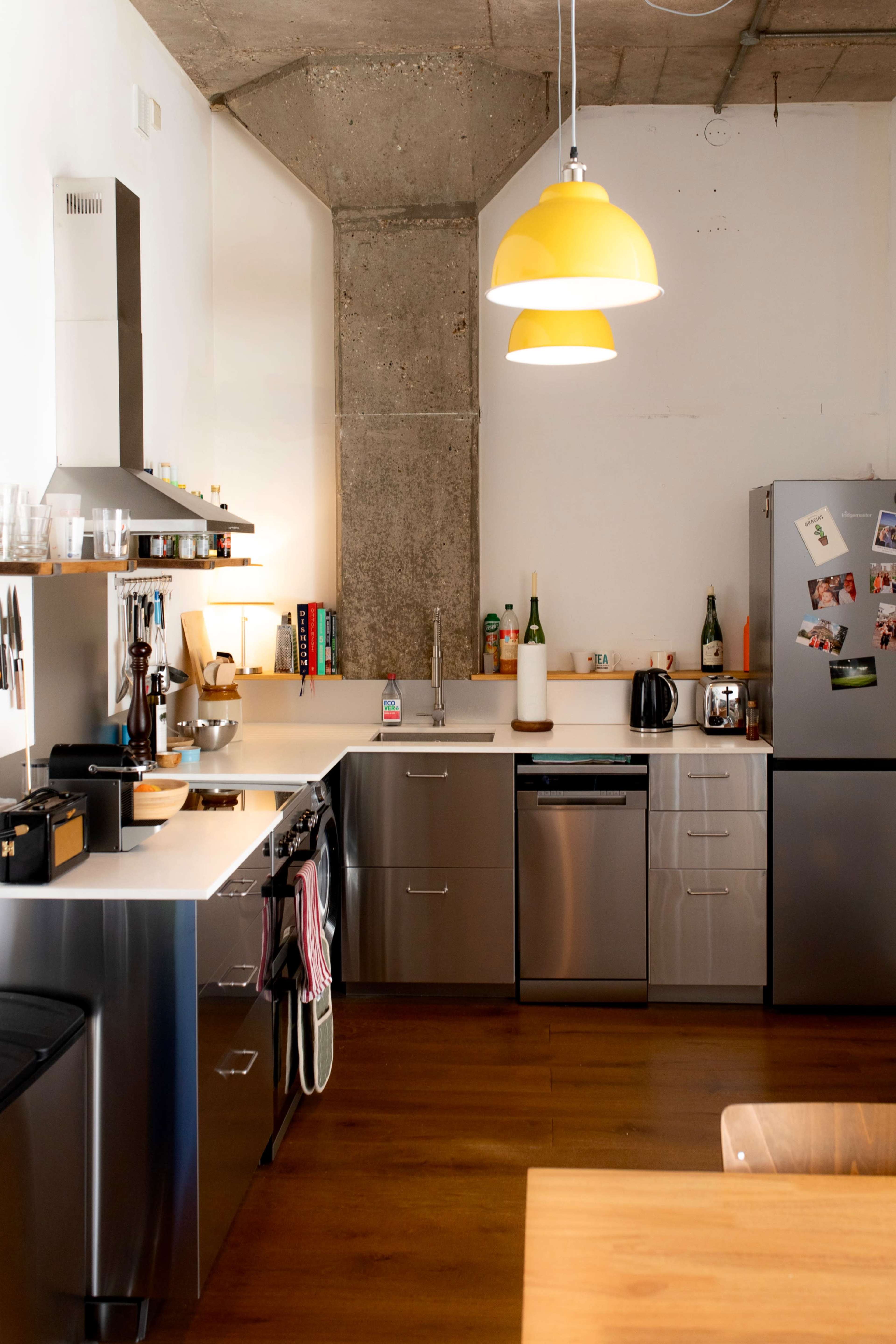 The kitchen features stainless steel appliances, wooden flooring, and a yellow pendant light hanging above a countertop with various utensils and a sink.