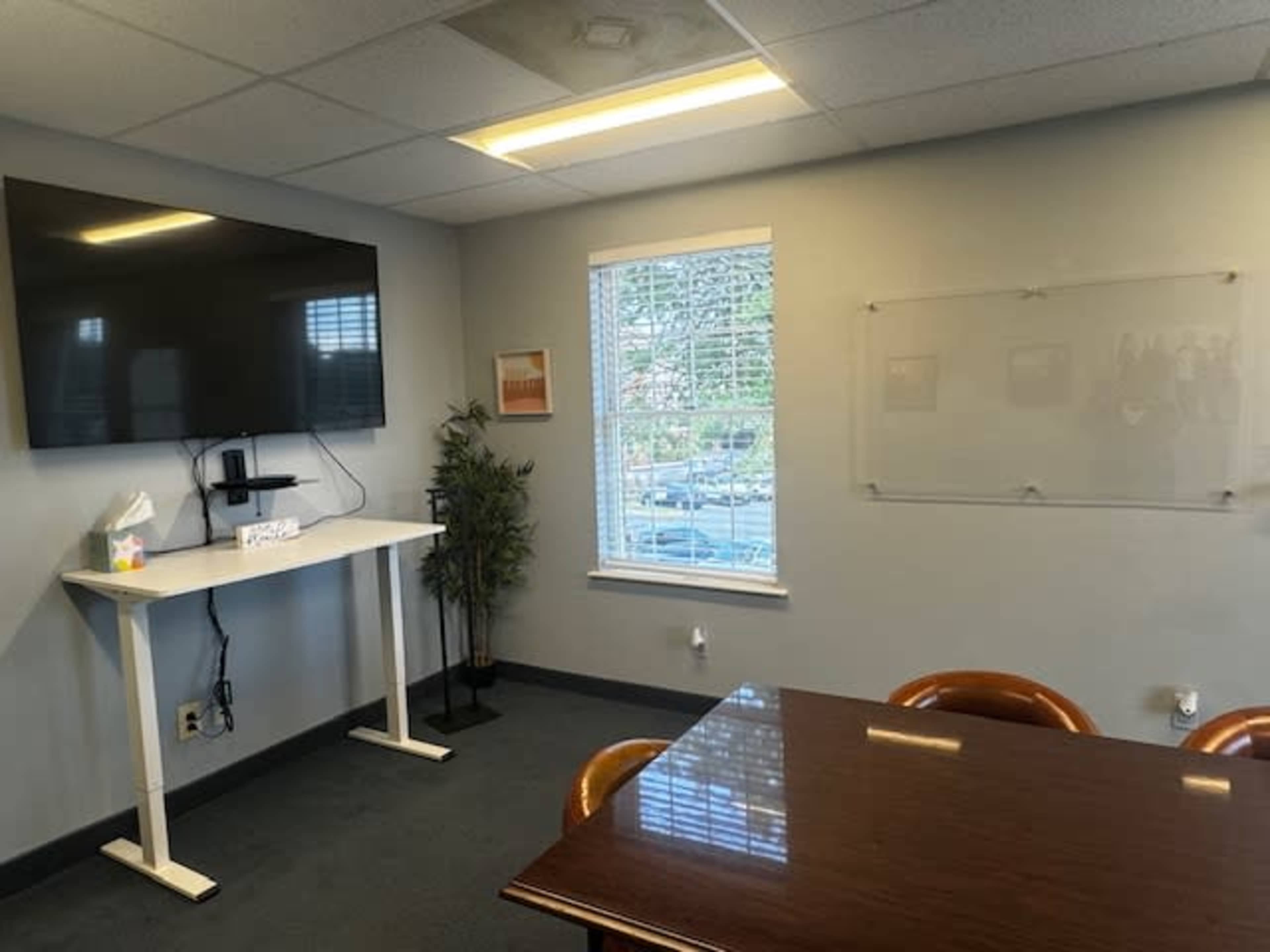 A modern office meeting room with a large wall-mounted TV, a standing desk, a potted plant, and a wooden conference table.