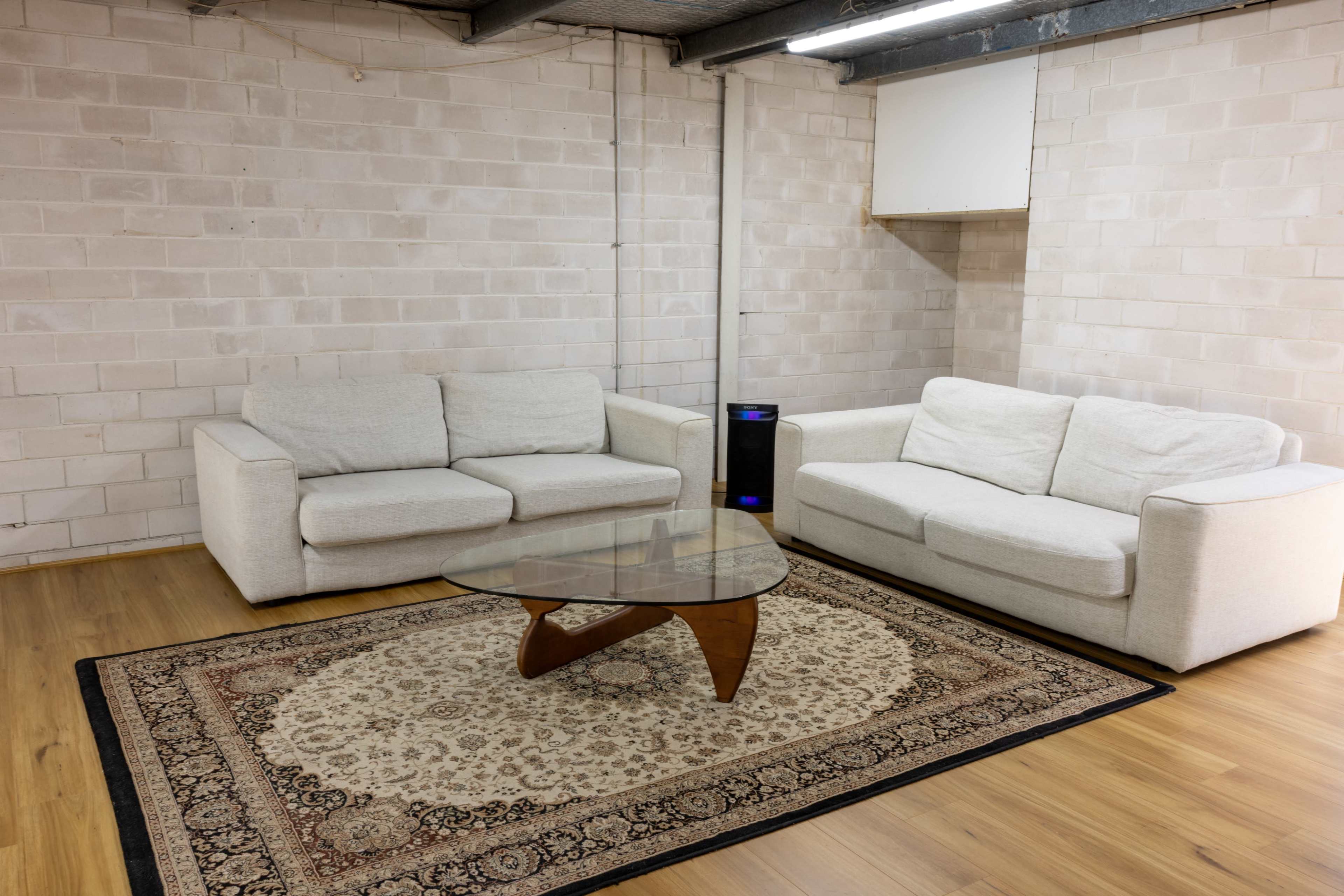 A furnished room with two light-colored sofas, a glass coffee table, and a patterned rug on wooden flooring against a brick wall.