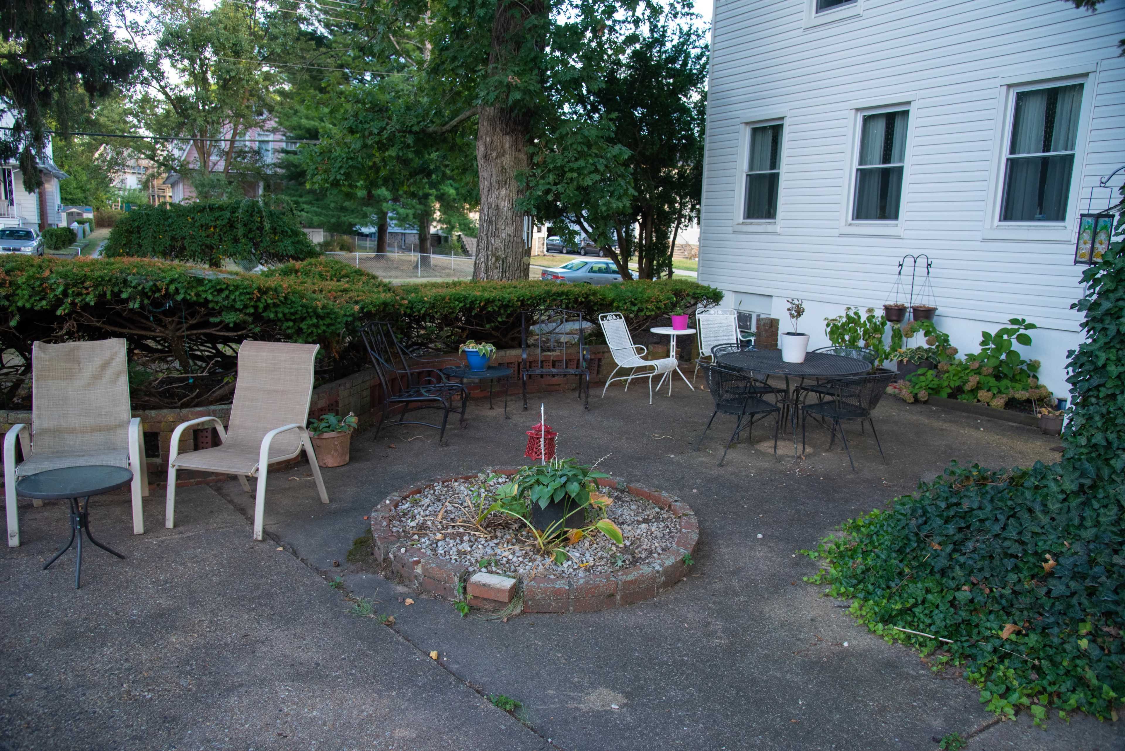 The image shows an outdoor patio area with various seating options, a round garden bed, and greenery surrounding it.