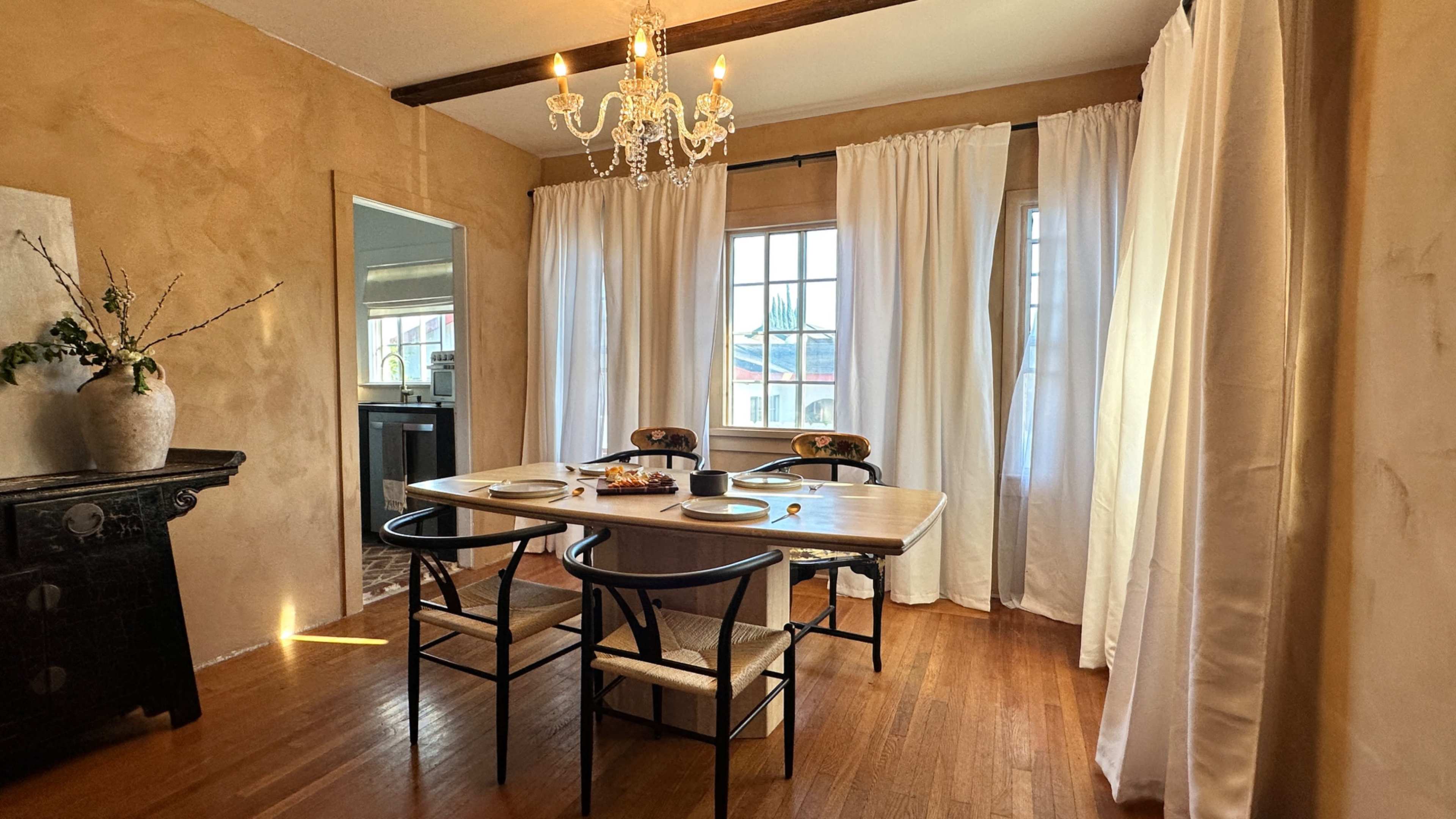 A dining room features a wooden table set for four, surrounded by chairs, with a chandelier above and large window draped with white curtains.