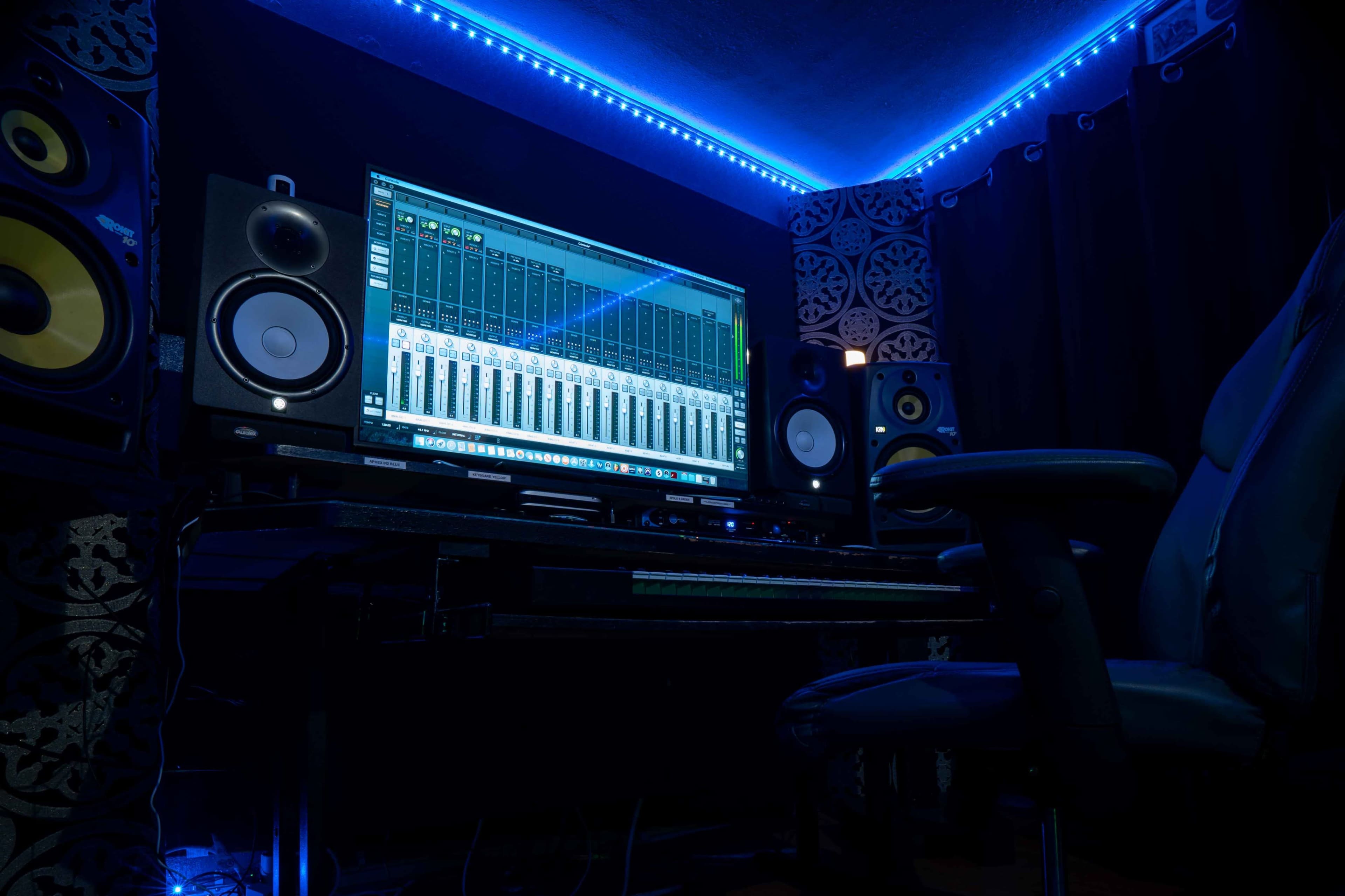 A home recording studio features a computer screen displaying audio mixer software, flanked by studio monitors and illuminated by blue LED lights.