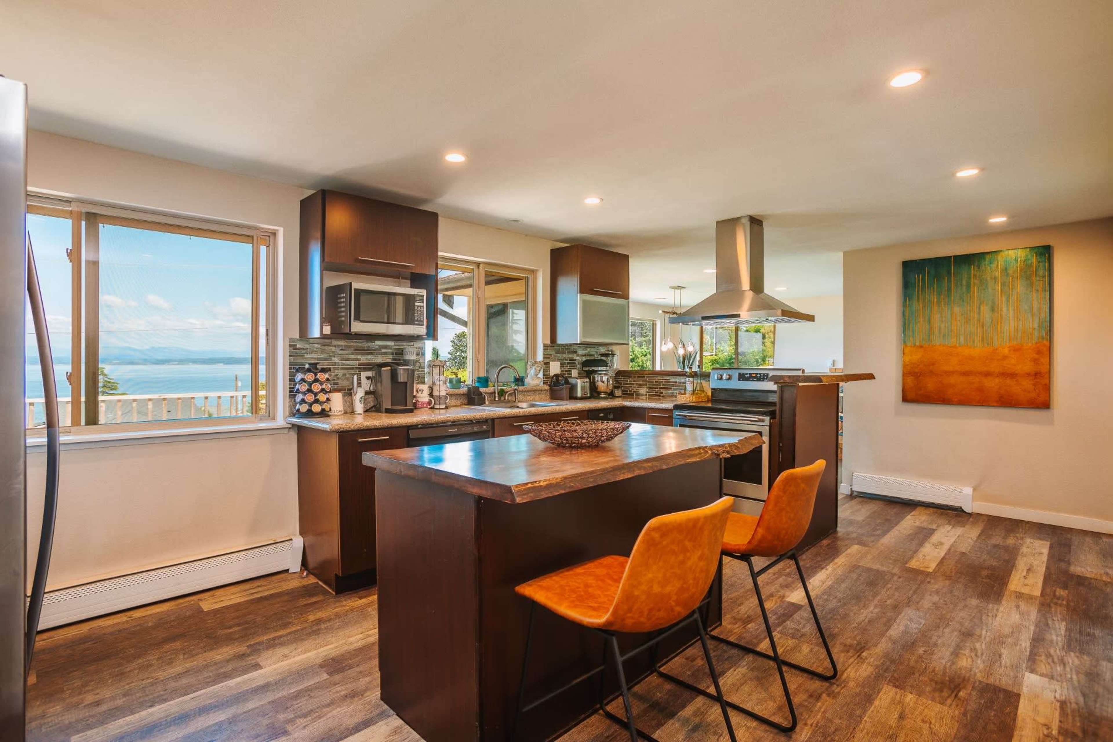 A modern kitchen with wooden cabinetry, a central island featuring two yellow chairs, and large windows offering a view of a body of water.