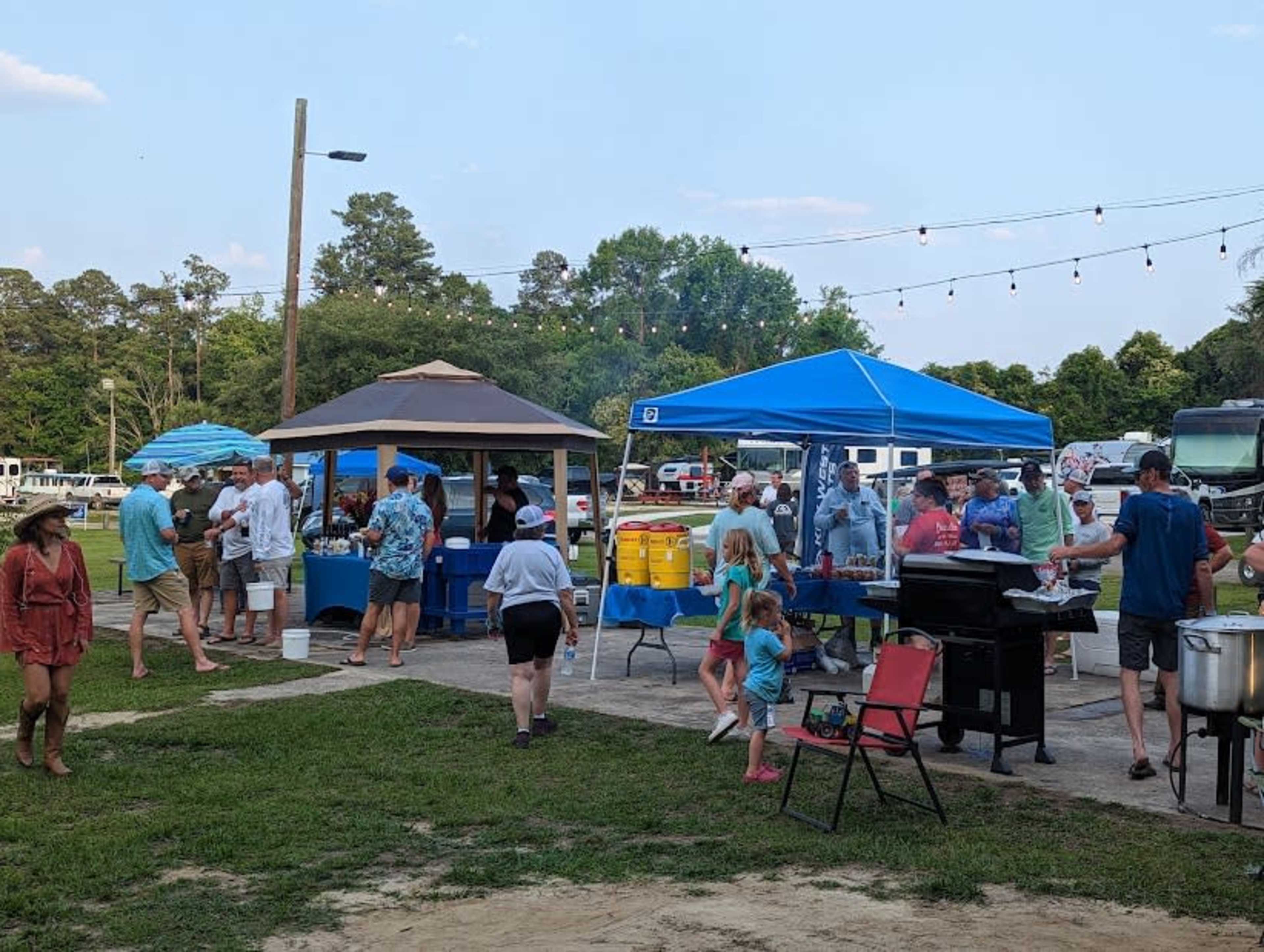 A gathering of people is taking place outdoors with tents, tables, and food preparations in a grassy area.