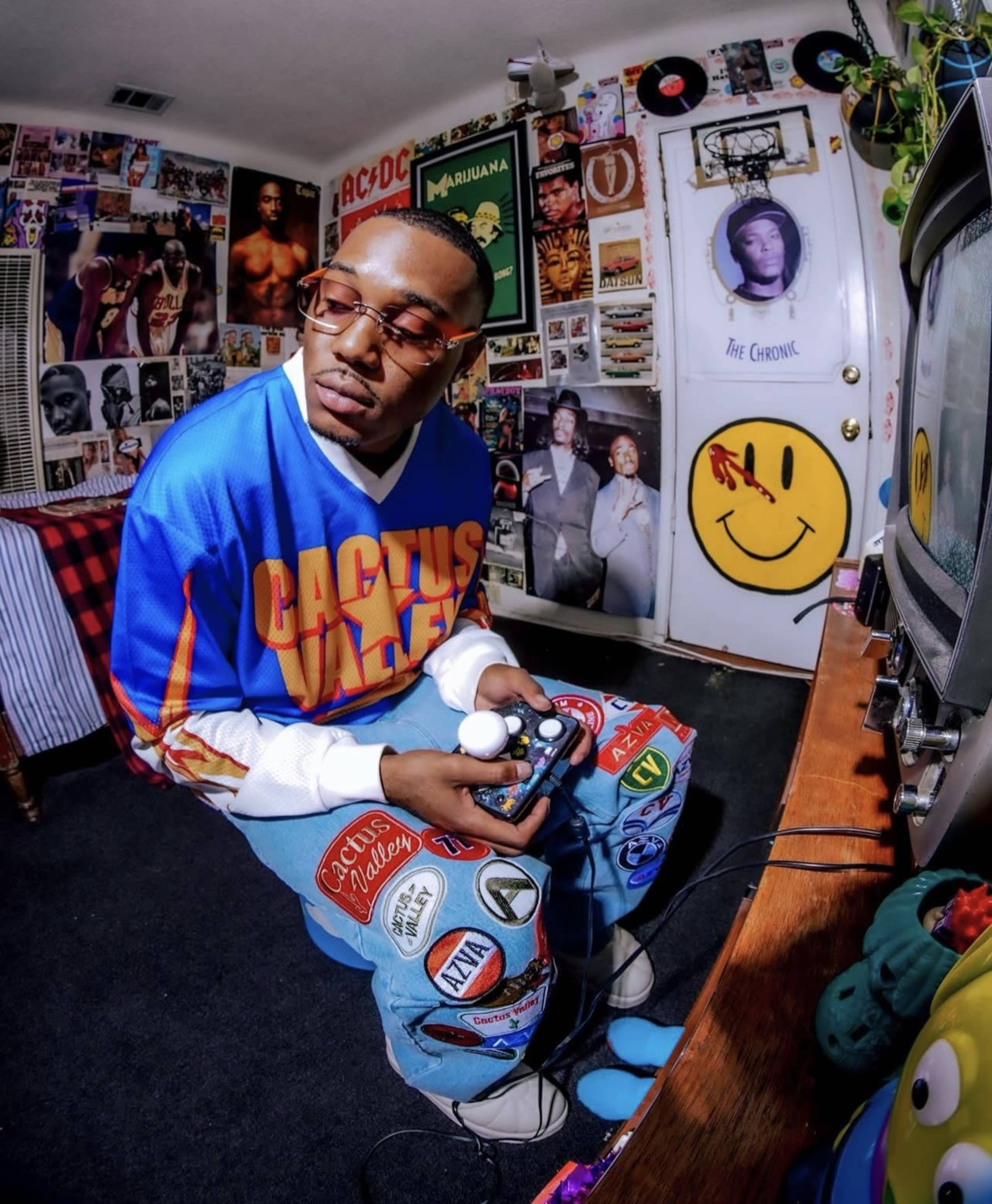 A person in a colorful retro jersey sits on the floor, holding a video game controller in front of a vintage television set surrounded by posters and memorabilia.
