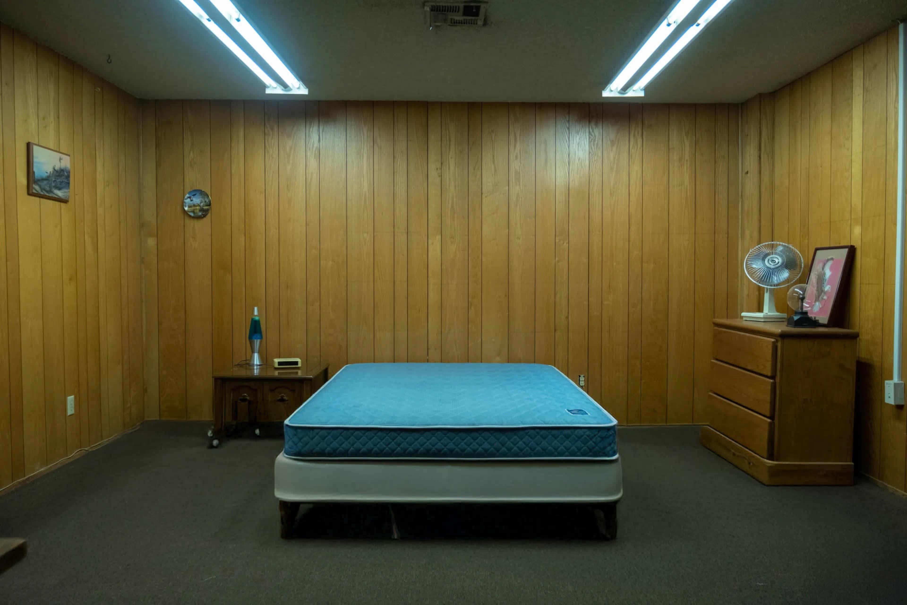 The image shows a sparsely furnished room with wooden paneling, featuring a blue mattress on a bed frame, a dresser, a bedside table, and a fan.