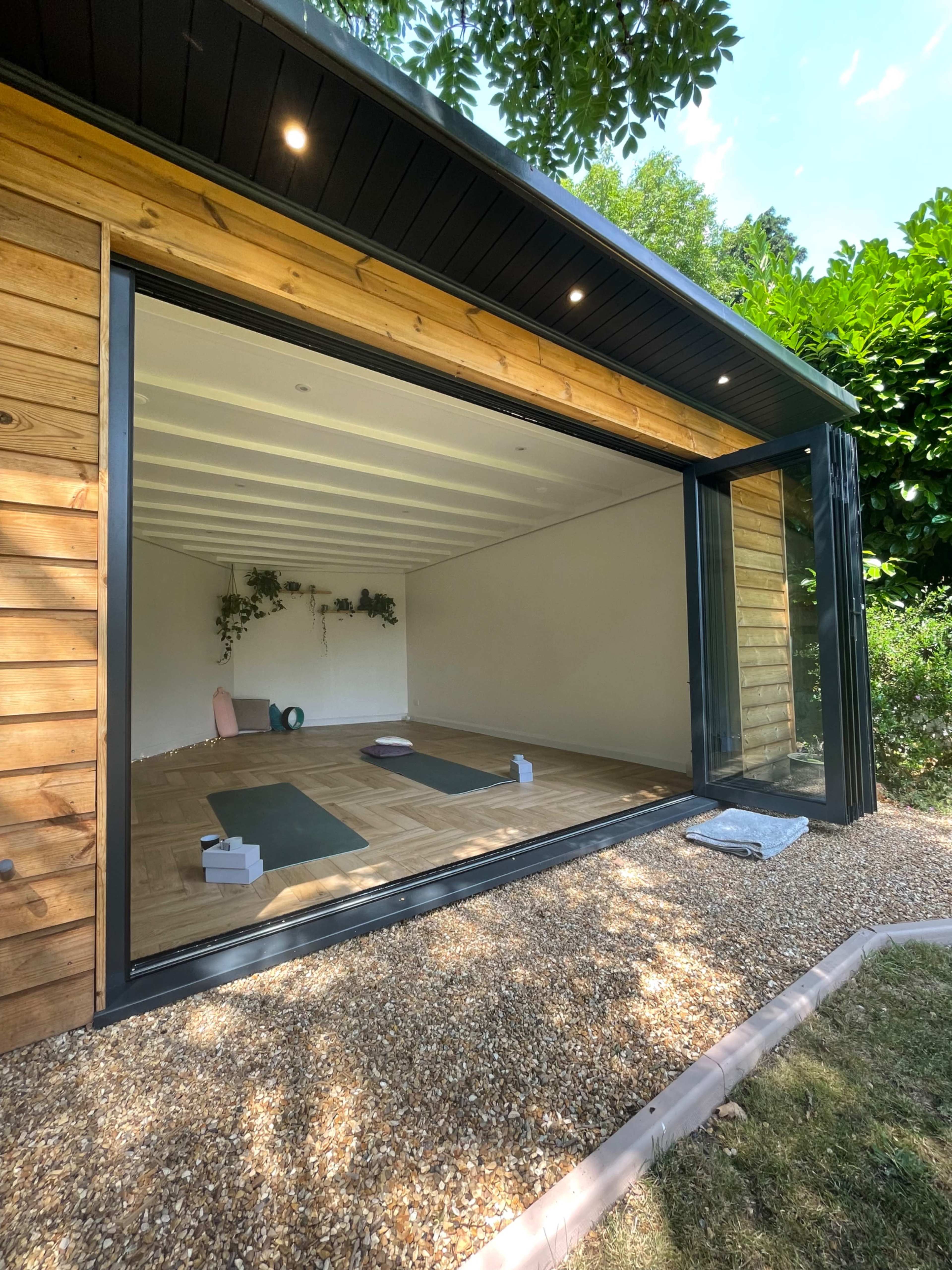 The image shows a spacious room with large glass doors that open to a gravel outdoor area, featuring wooden walls and minimalistic decor suitable for yoga or meditation.
