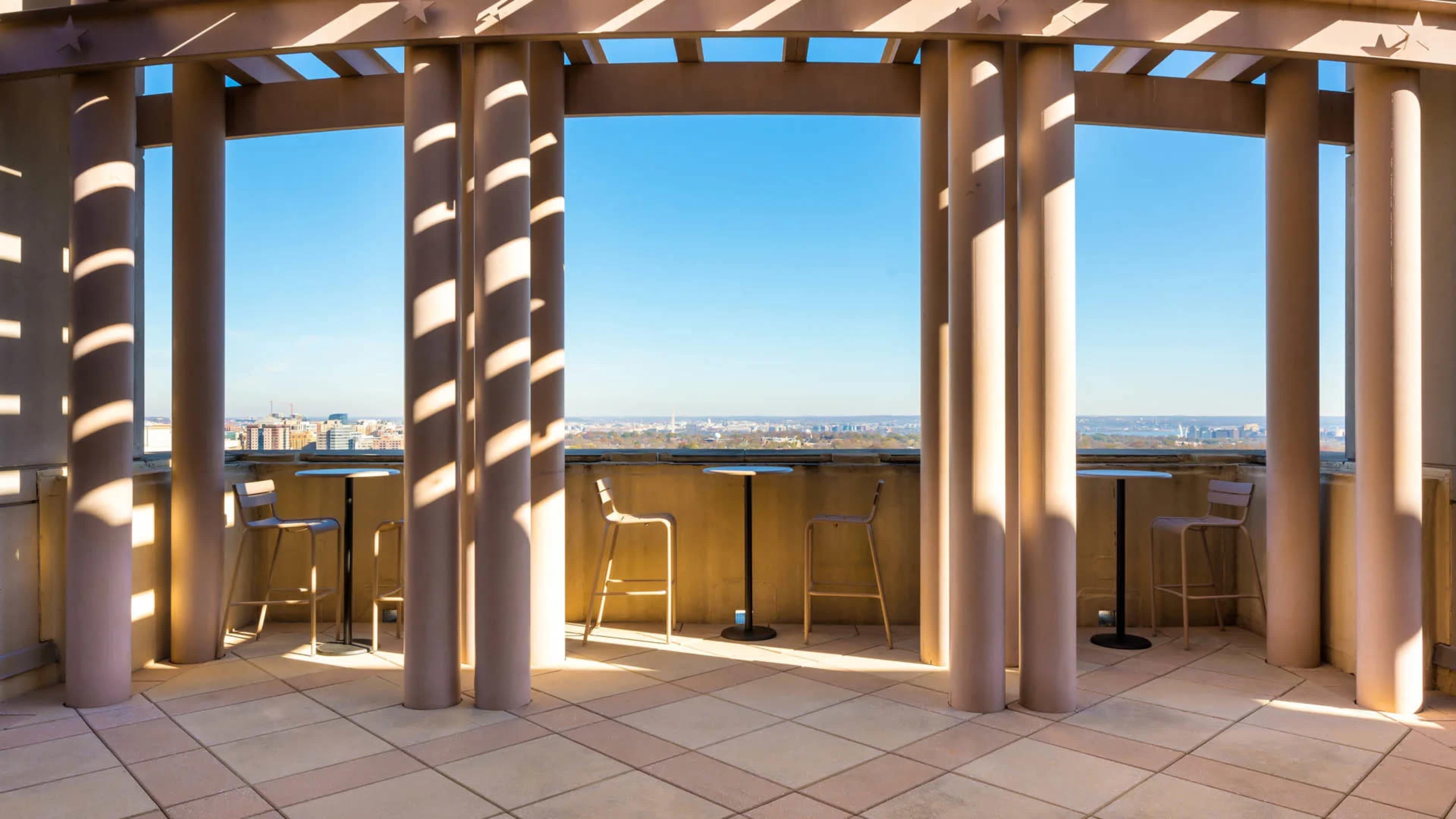 A rooftop bar area with tall pillars and high stools overlooking a cityscape under a clear blue sky.