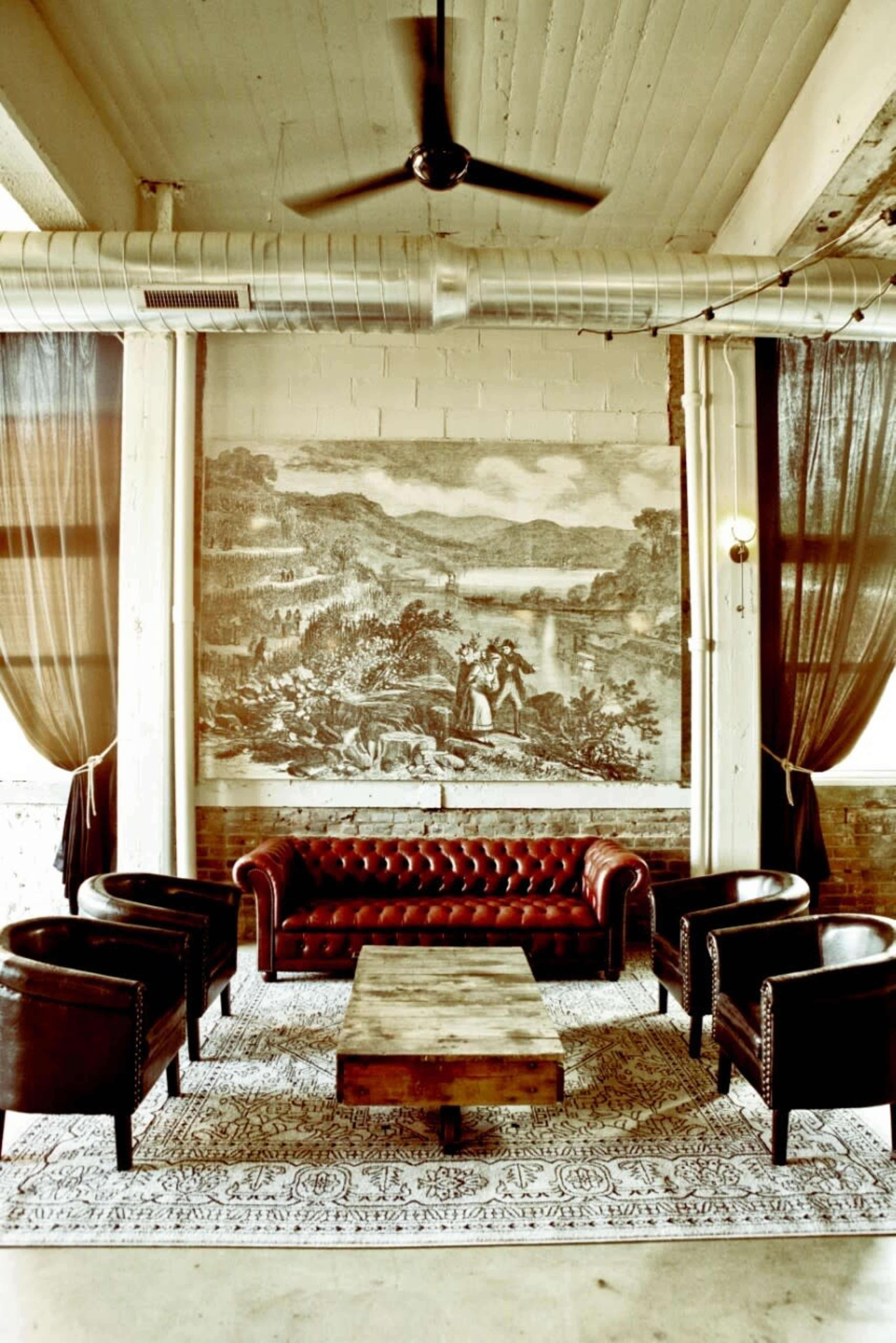 The image shows a stylish interior featuring a leather chesterfield sofa, four black armchairs, and a wooden coffee table, with a large mural depicting a landscape on the wall behind.