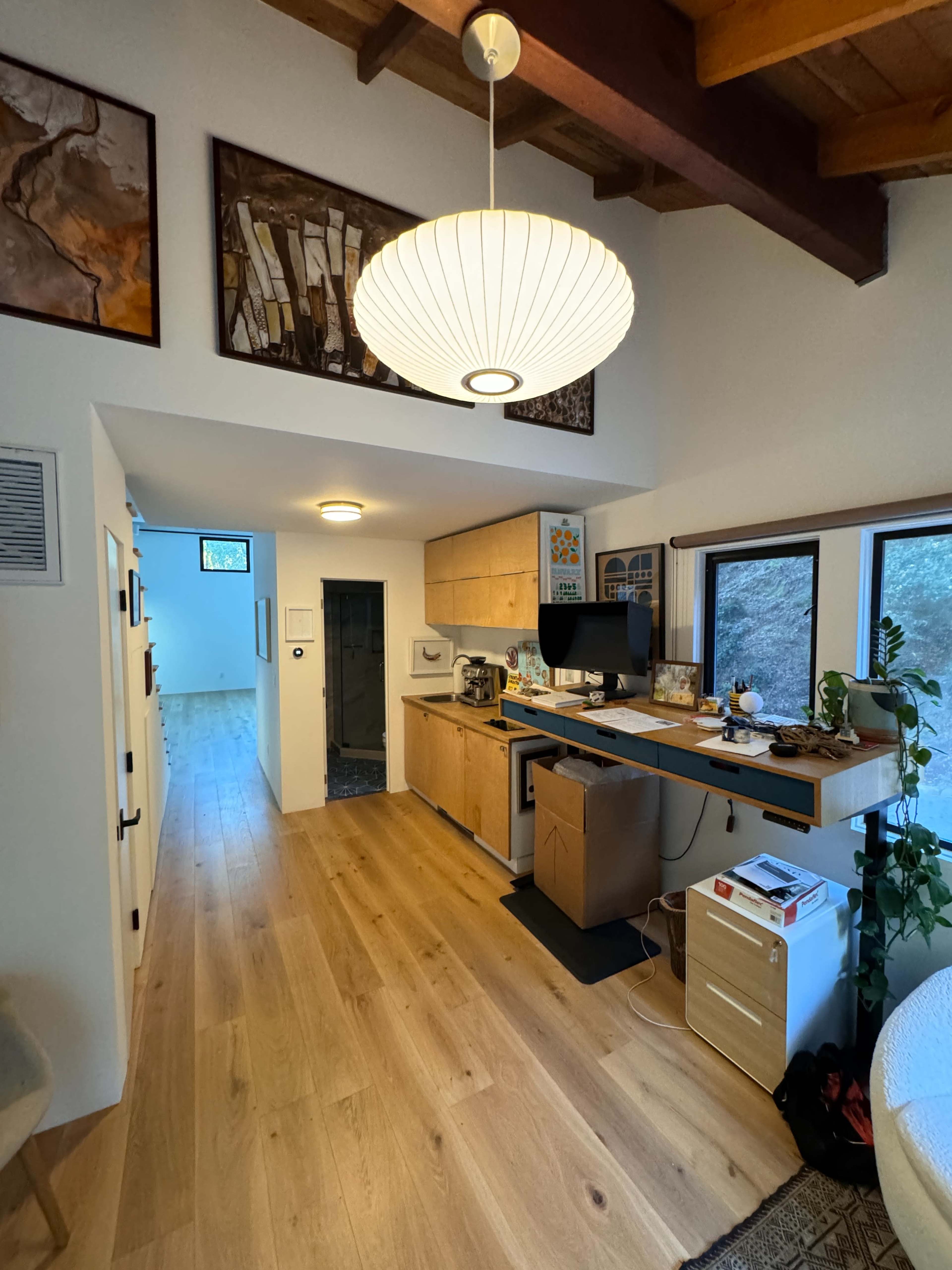A modern home office area featuring a wooden desk, a computer setup, and a large pendant light, with a hallway and windows visible in the background.