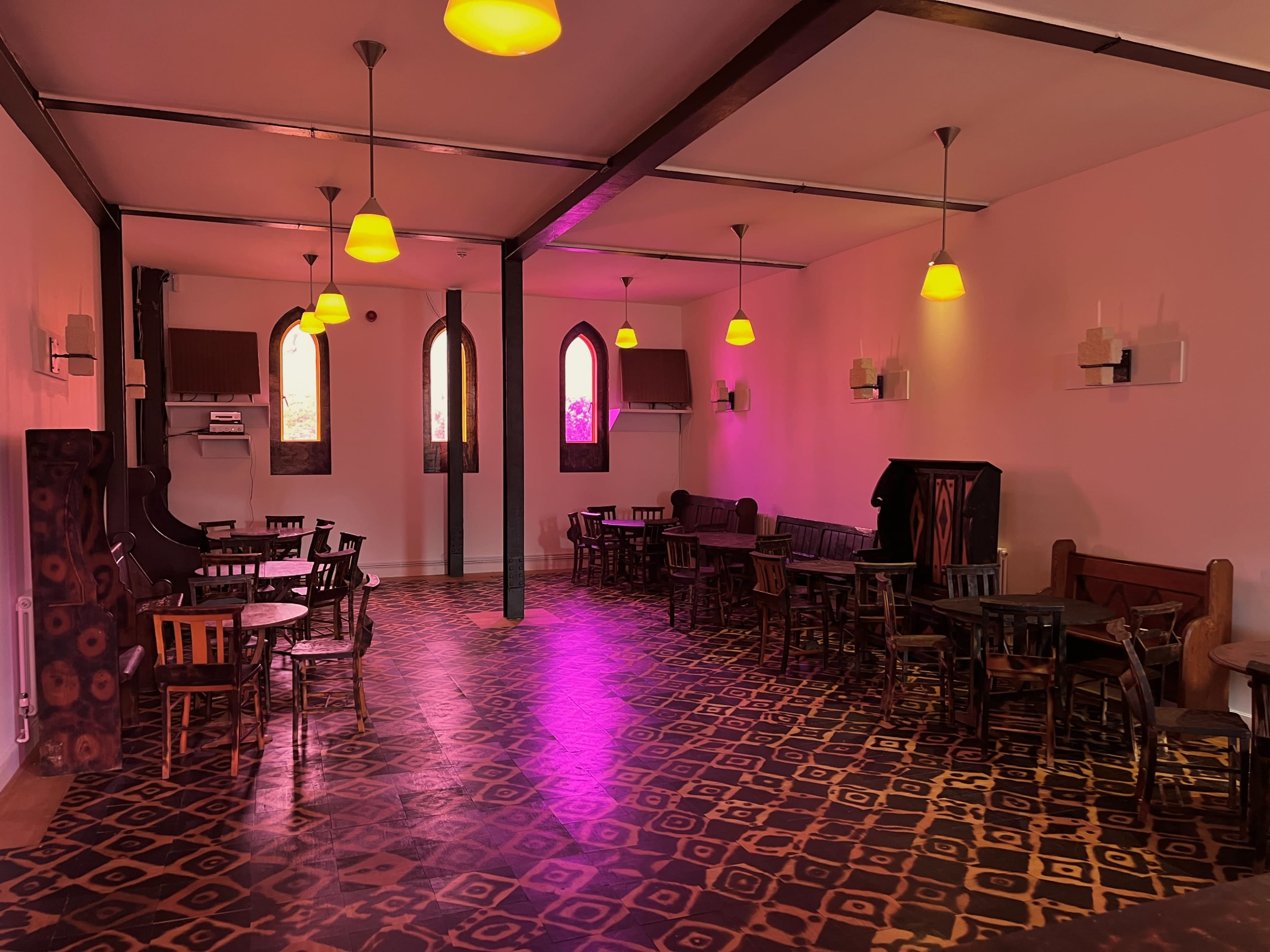 The image shows an empty interior of a café or restaurant featuring wooden tables and chairs, with pink walls and unique patterned flooring illuminated by warm light fixtures.