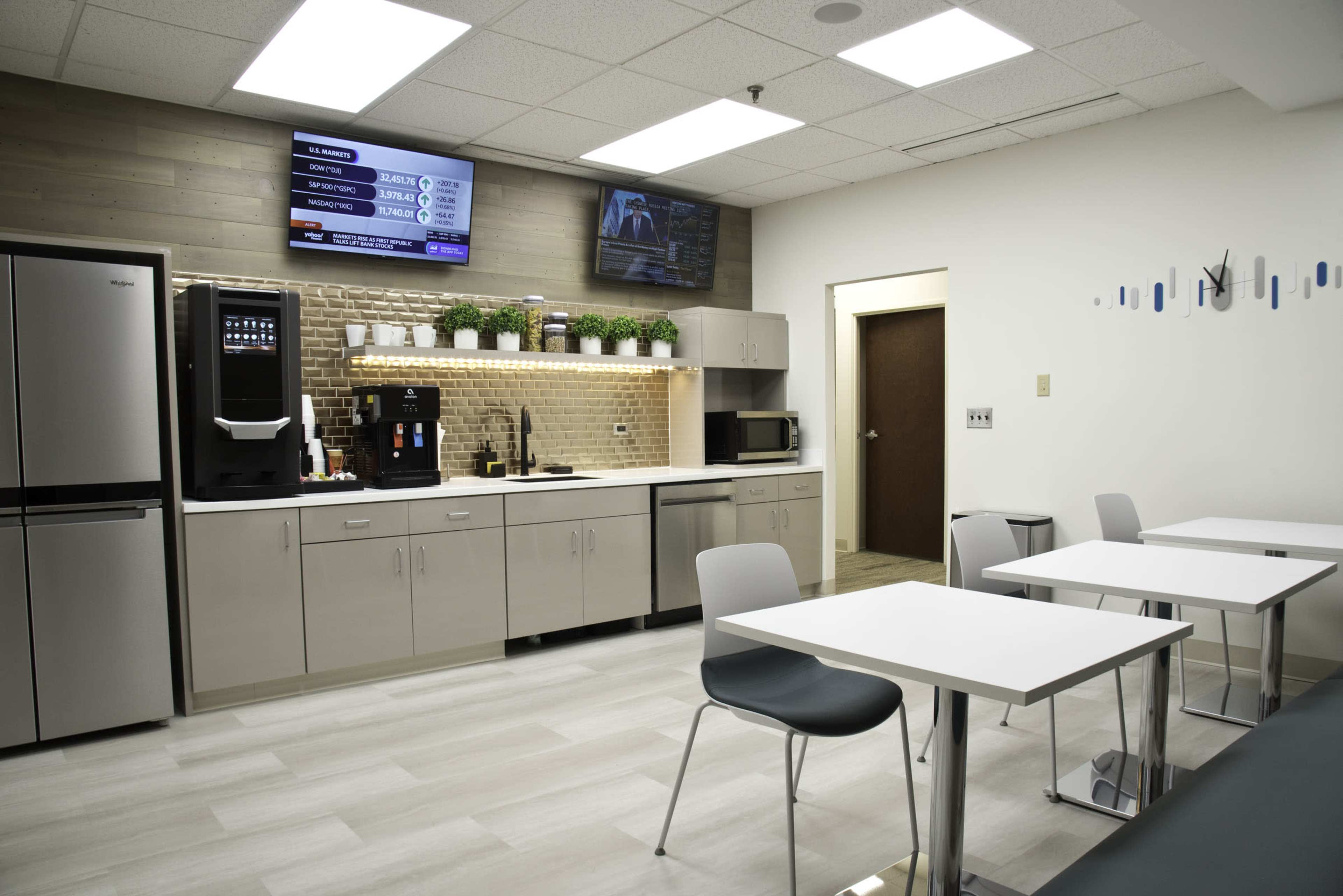 The image shows a modern break room with a kitchenette, featuring stainless steel appliances, white tables, and gray chairs.