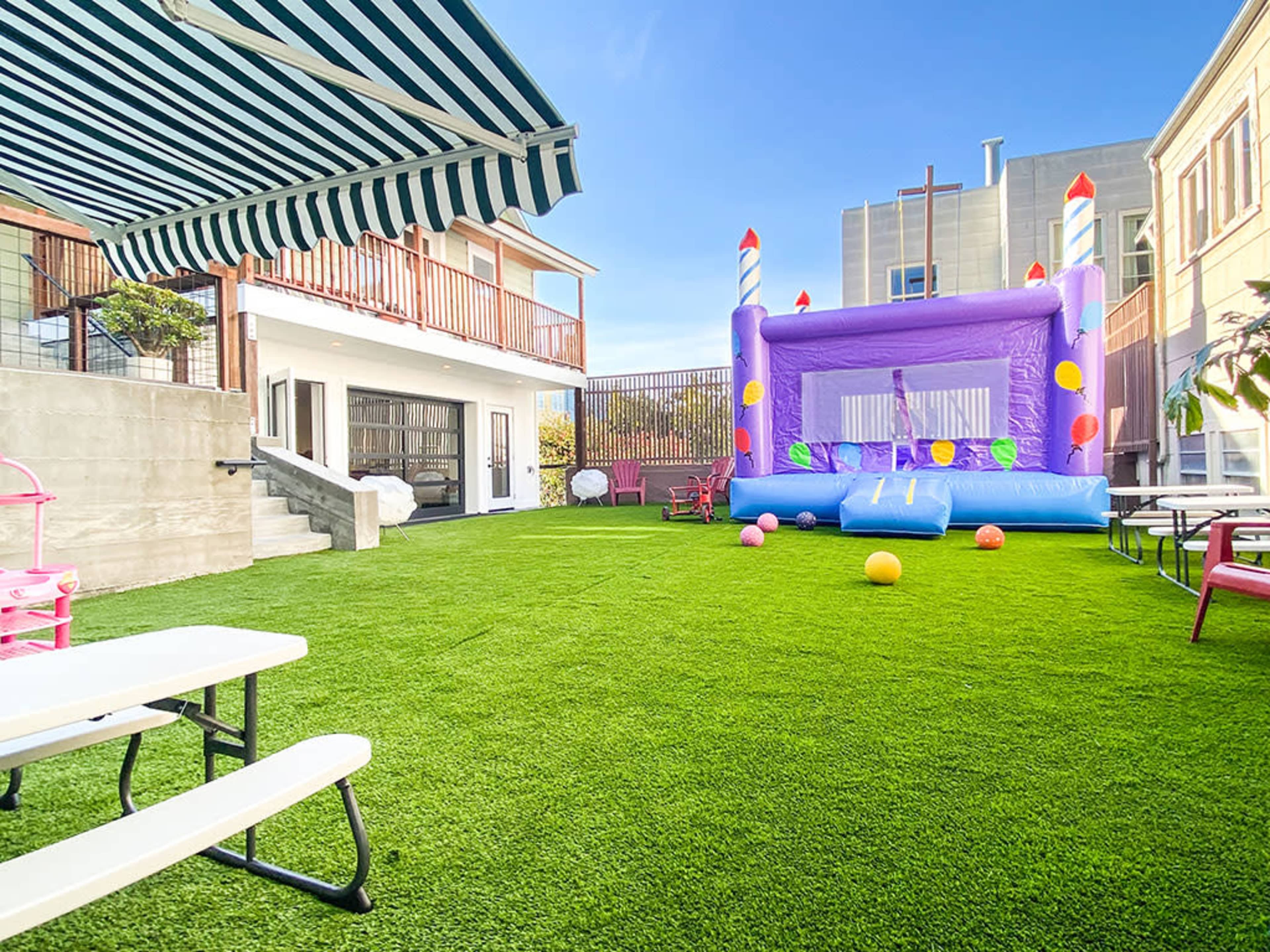 A colorful bounce house is set up on a green turf lawn next to a building with a shaded seating area.