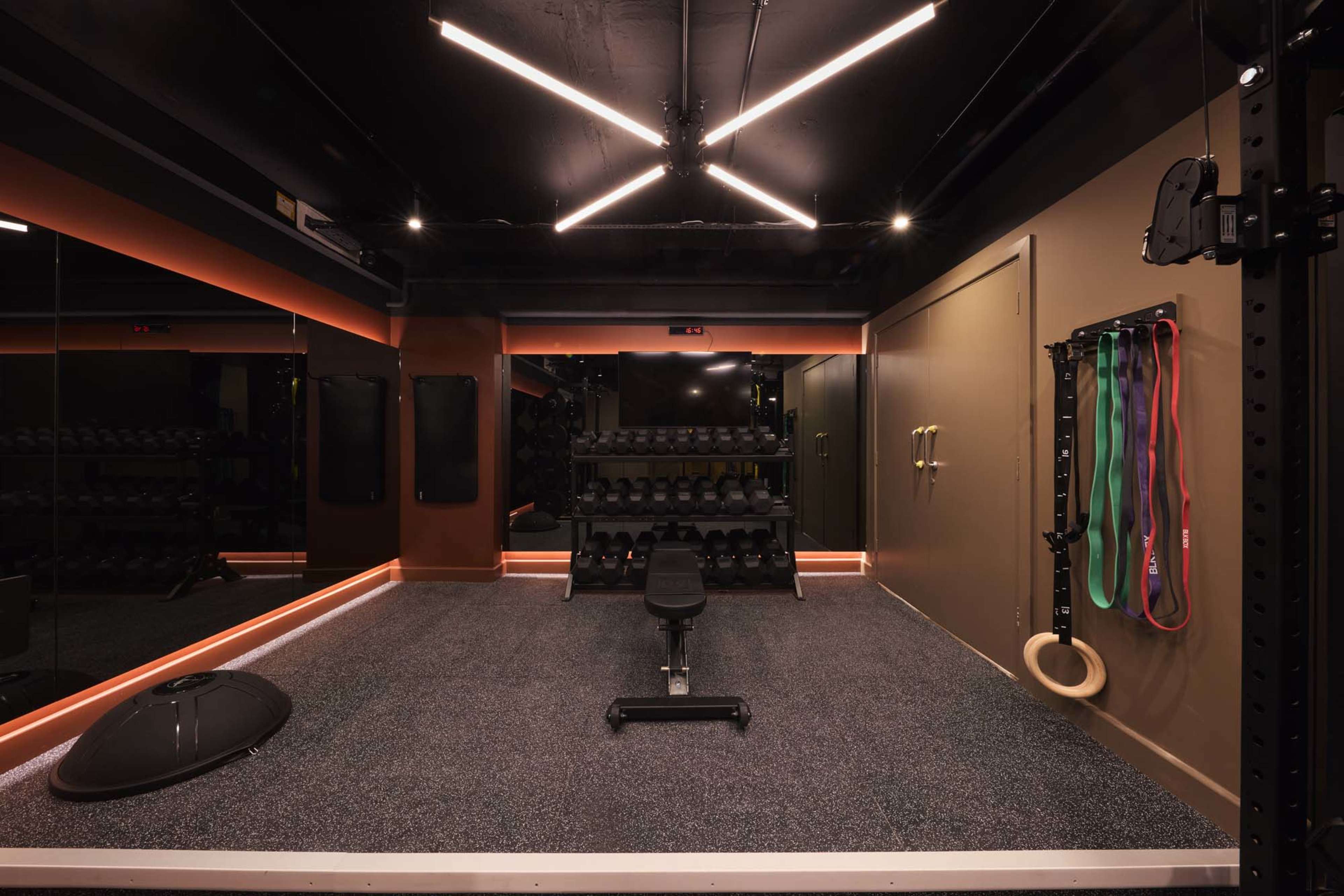 The image shows a modern home gym equipped with weights, a bench, and resistance bands, featuring sleek lighting and a textured floor.