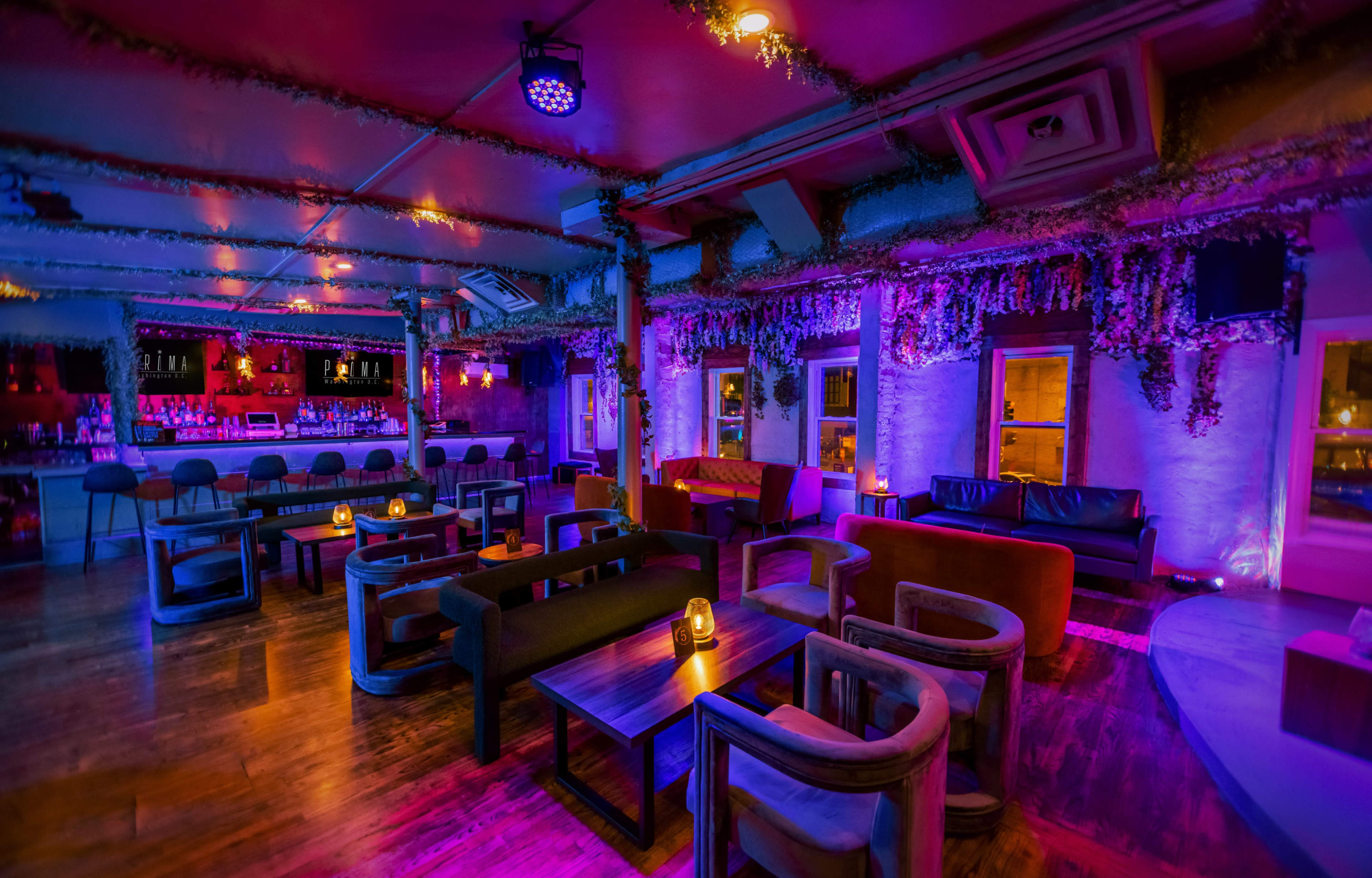 A vibrant bar interior with colorful lighting, featuring plush seating arrangements and a long bar area.