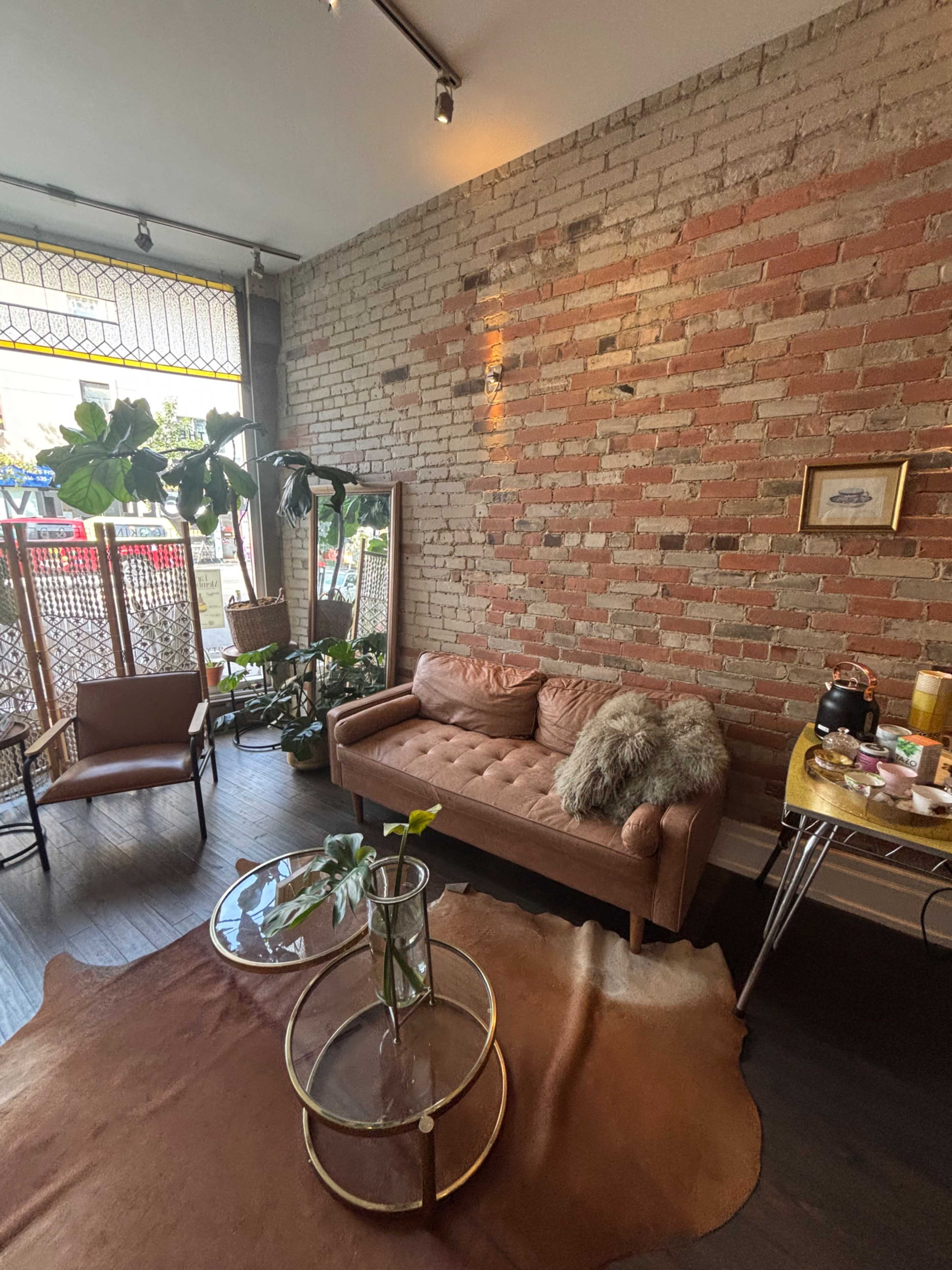 The scene depicts a cozy interior with a brick wall, a pink sofa, a round coffee table, and various plants, illuminated by natural light from a large window.