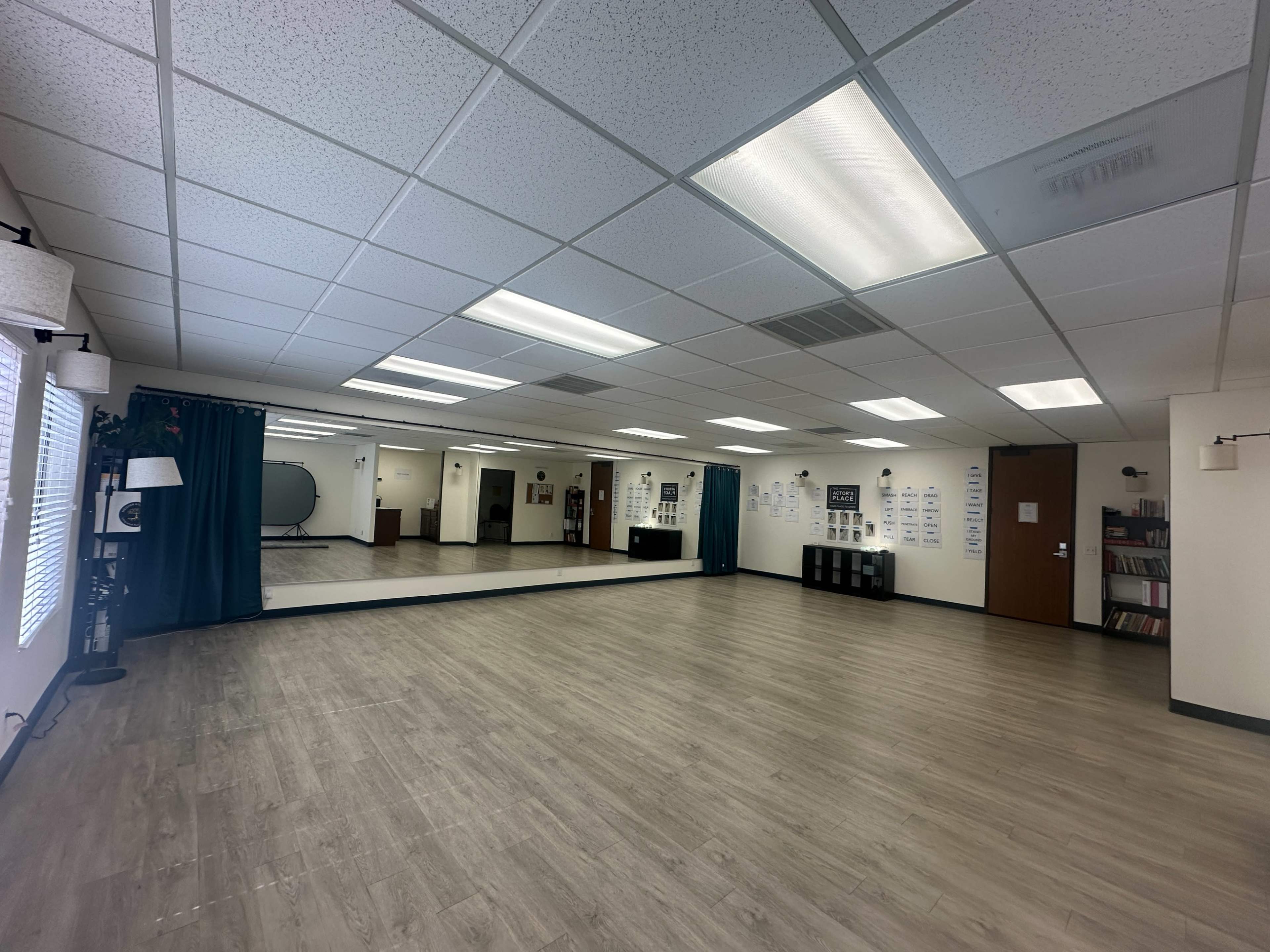 The image shows an empty dance studio with mirrors along one wall, wooden flooring, and large windows covered by curtains.
