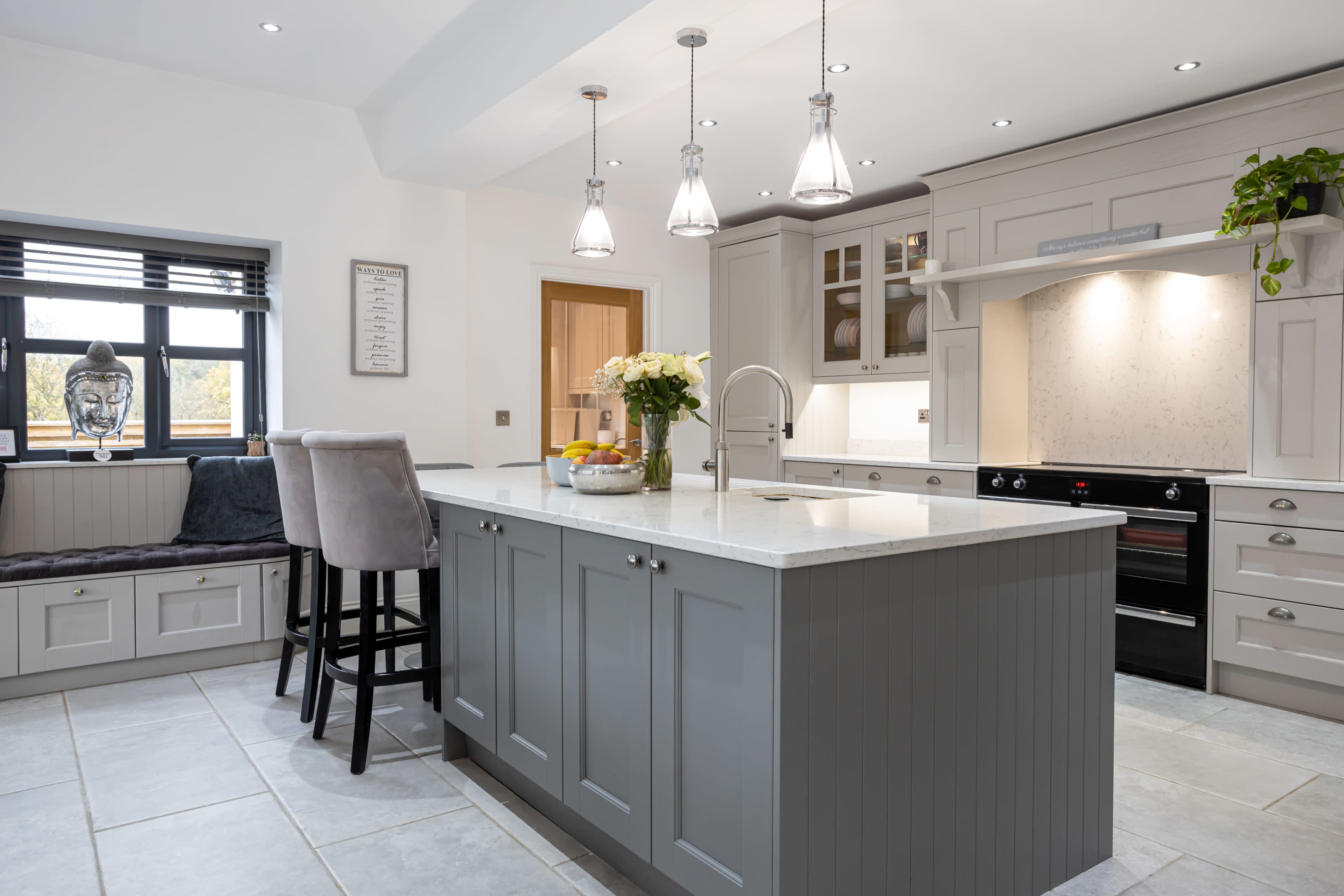 A modern kitchen features a central island with a light gray finish, bar stools, pendant lighting, and ample cabinetry, with a large window providing natural light.