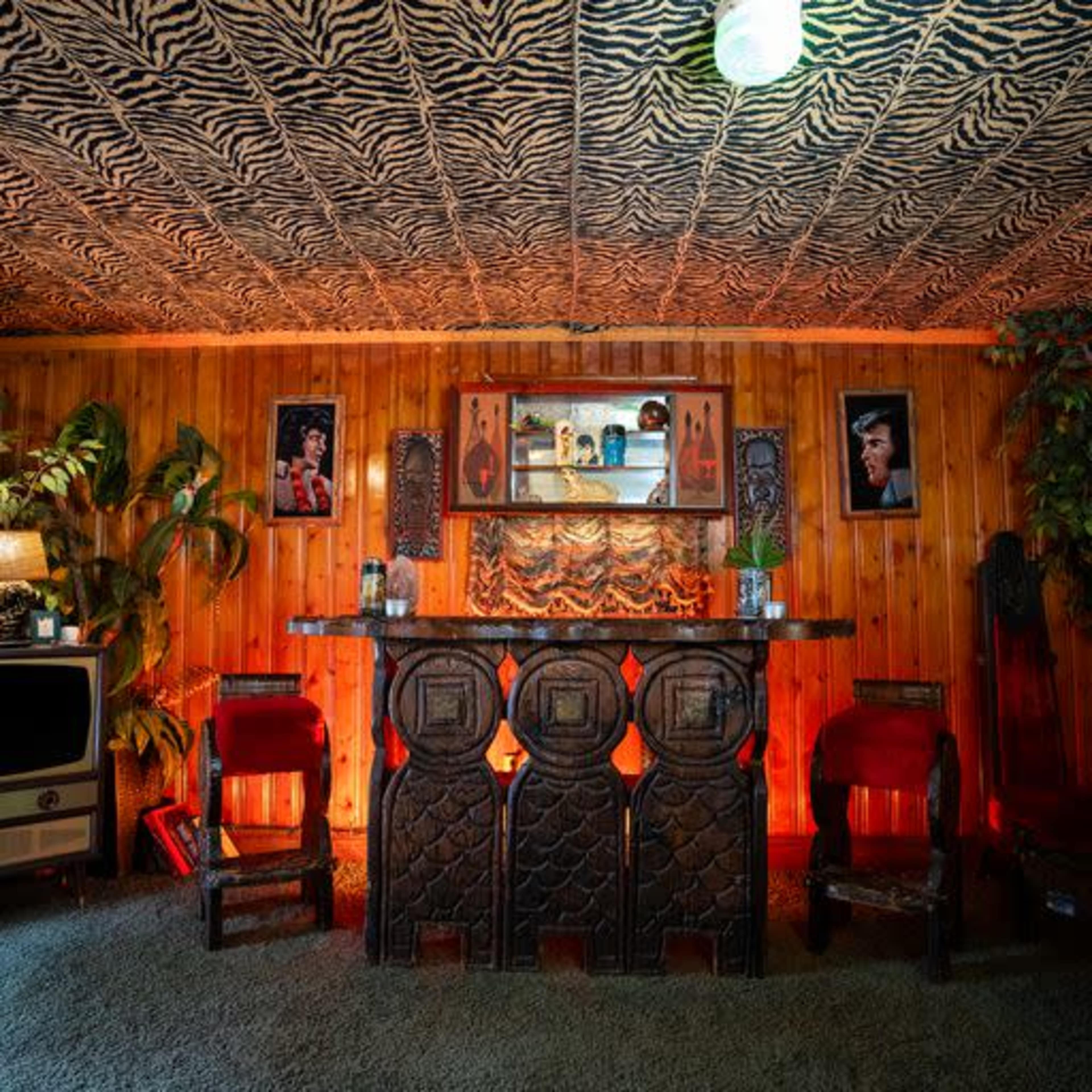 A retro room with a wooden panel wall, a zebra-print ceiling, a carved wooden bar, and two red chairs, surrounded by plants and illuminated by a hanging light.