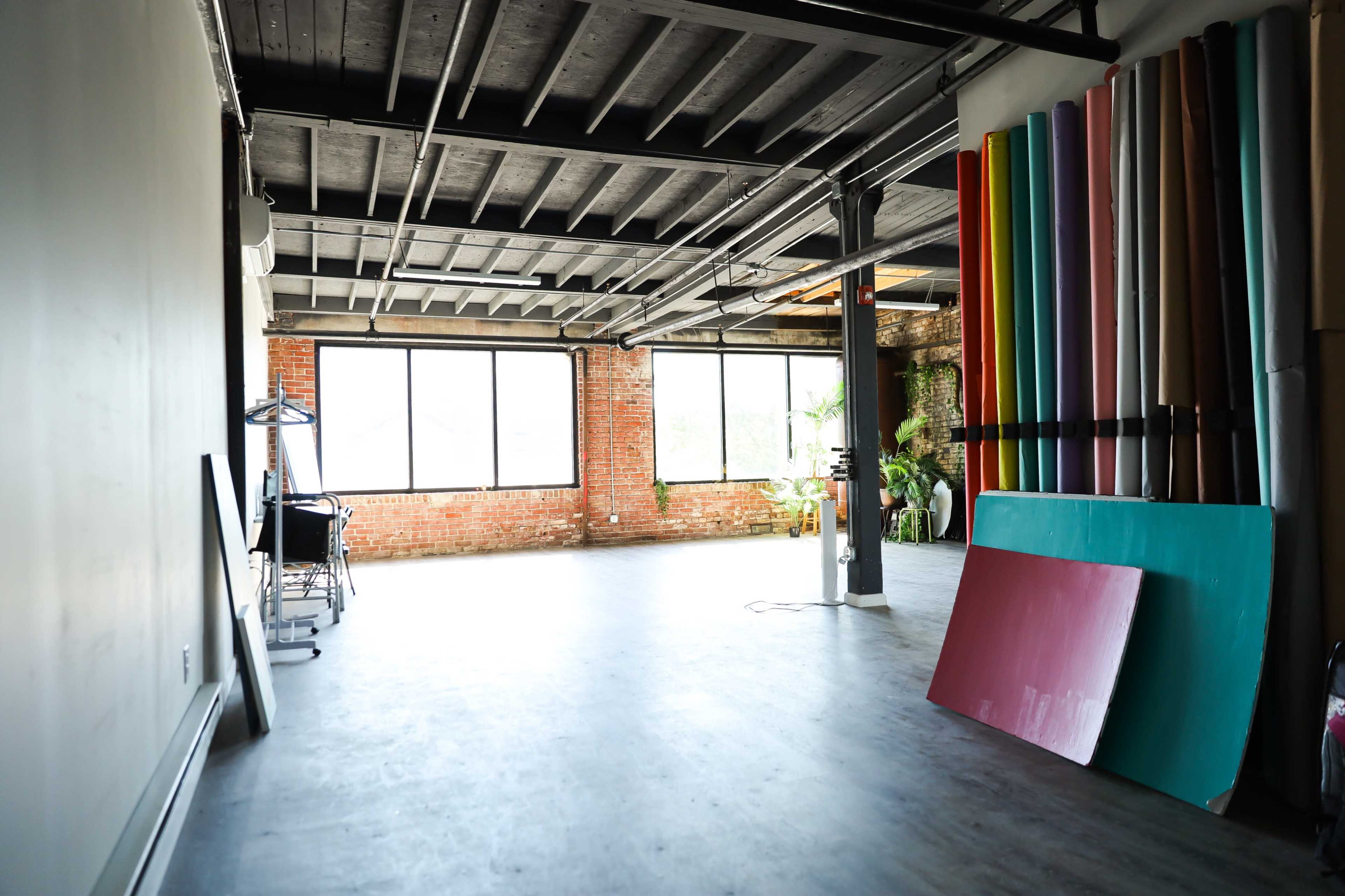 An empty studio space with large windows, exposed brick walls, and colorful panels stacked against one side.