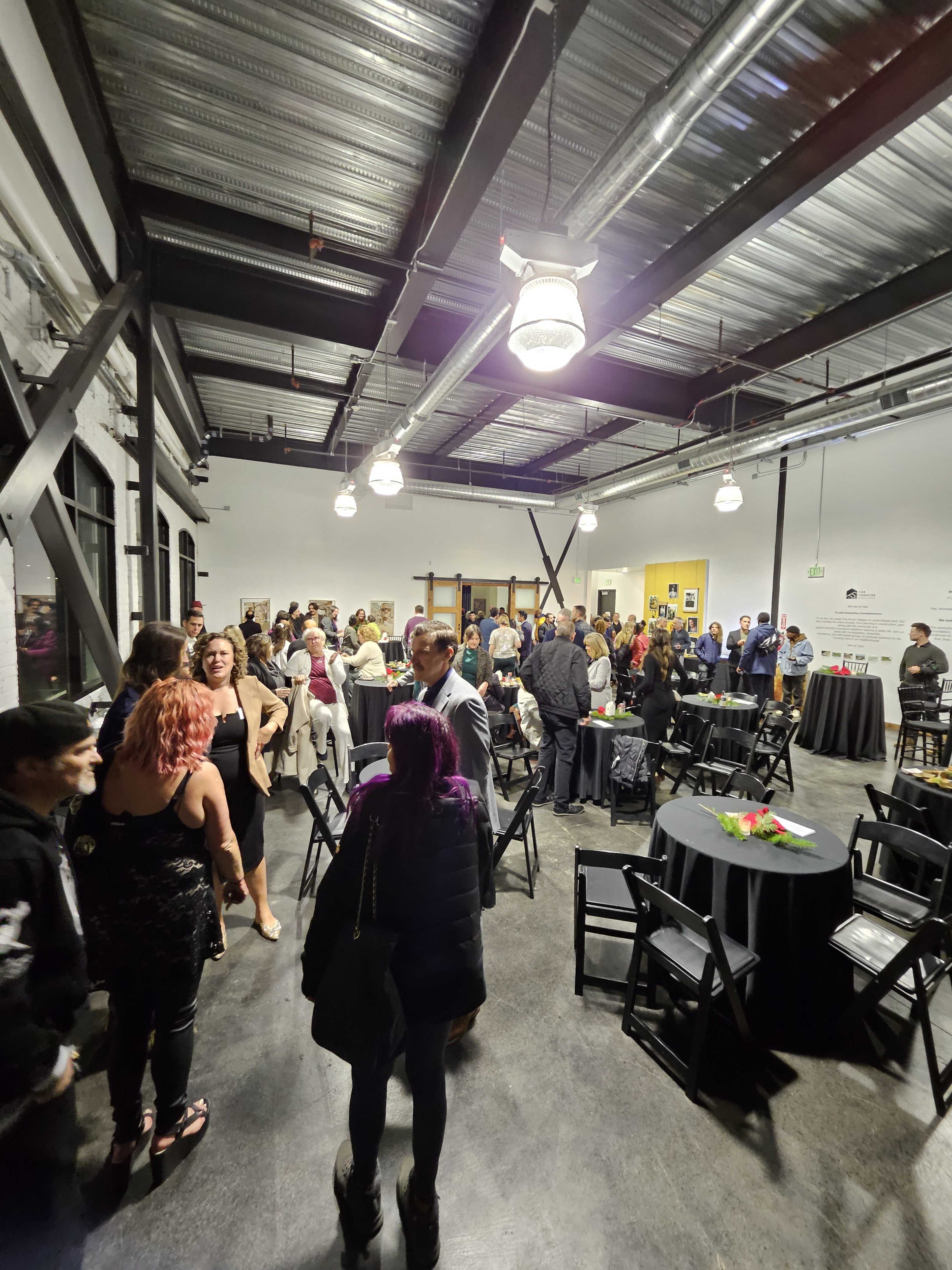 A crowd of people mingles in a large, industrial-style event space with black chairs and tables.