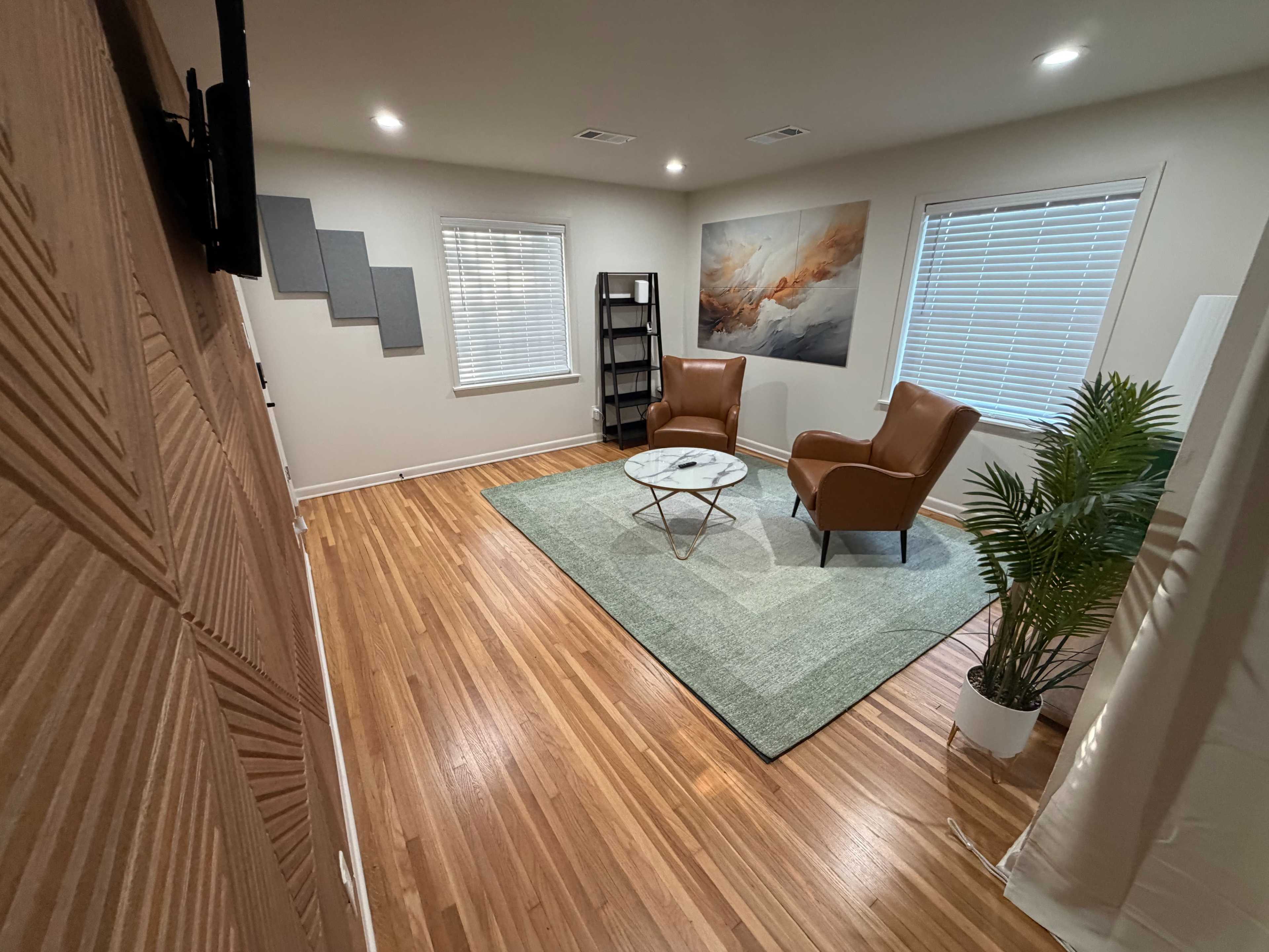 The image depicts a well-lit room featuring two brown armchairs, a round glass coffee table on a green area rug, a plant in the corner, and an abstract painting on the wall.