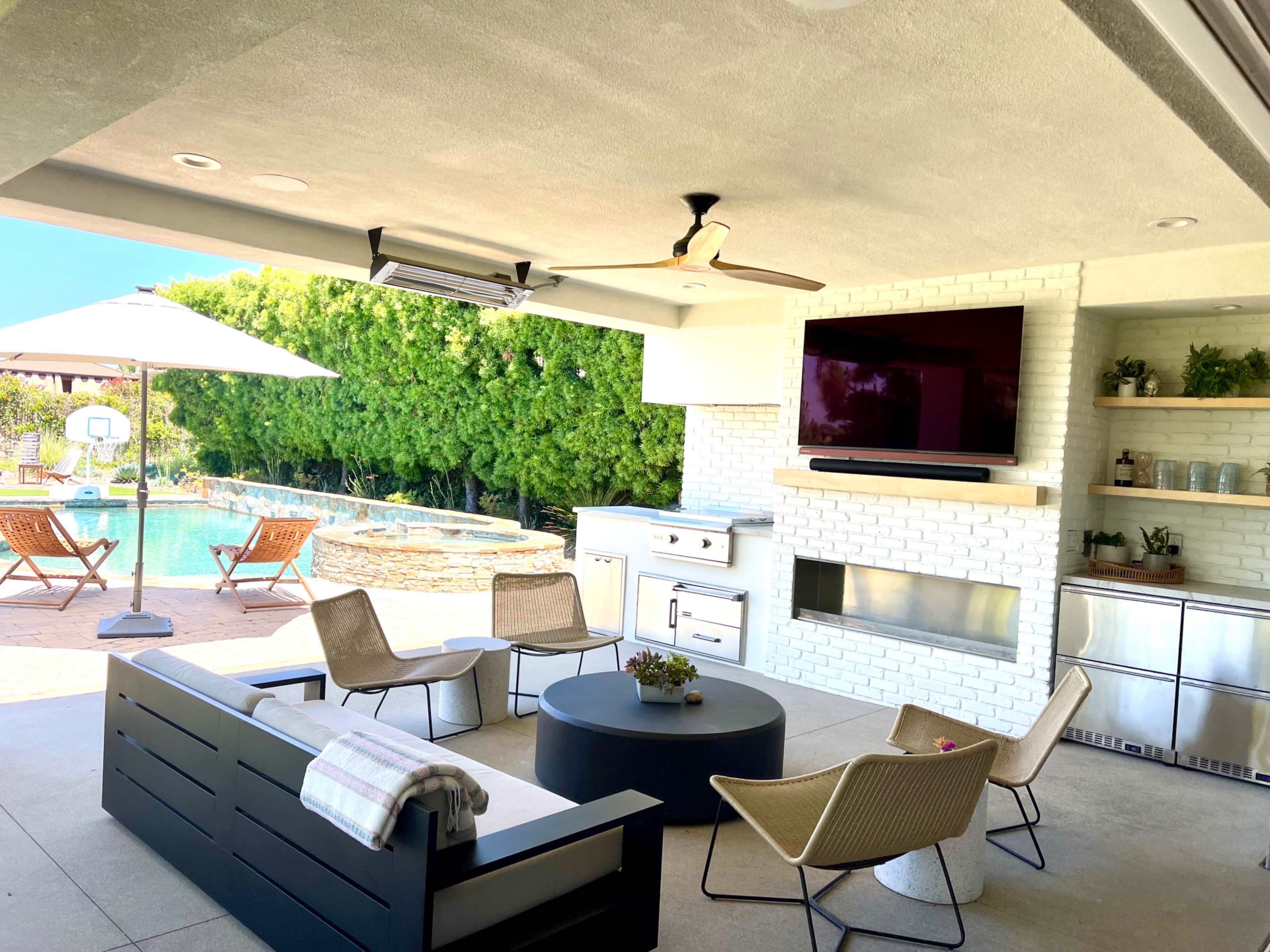 An outdoor patio features a seating area with modern furniture, a kitchen setup, a television mounted on the wall, and a view of a swimming pool and landscaped greenery.