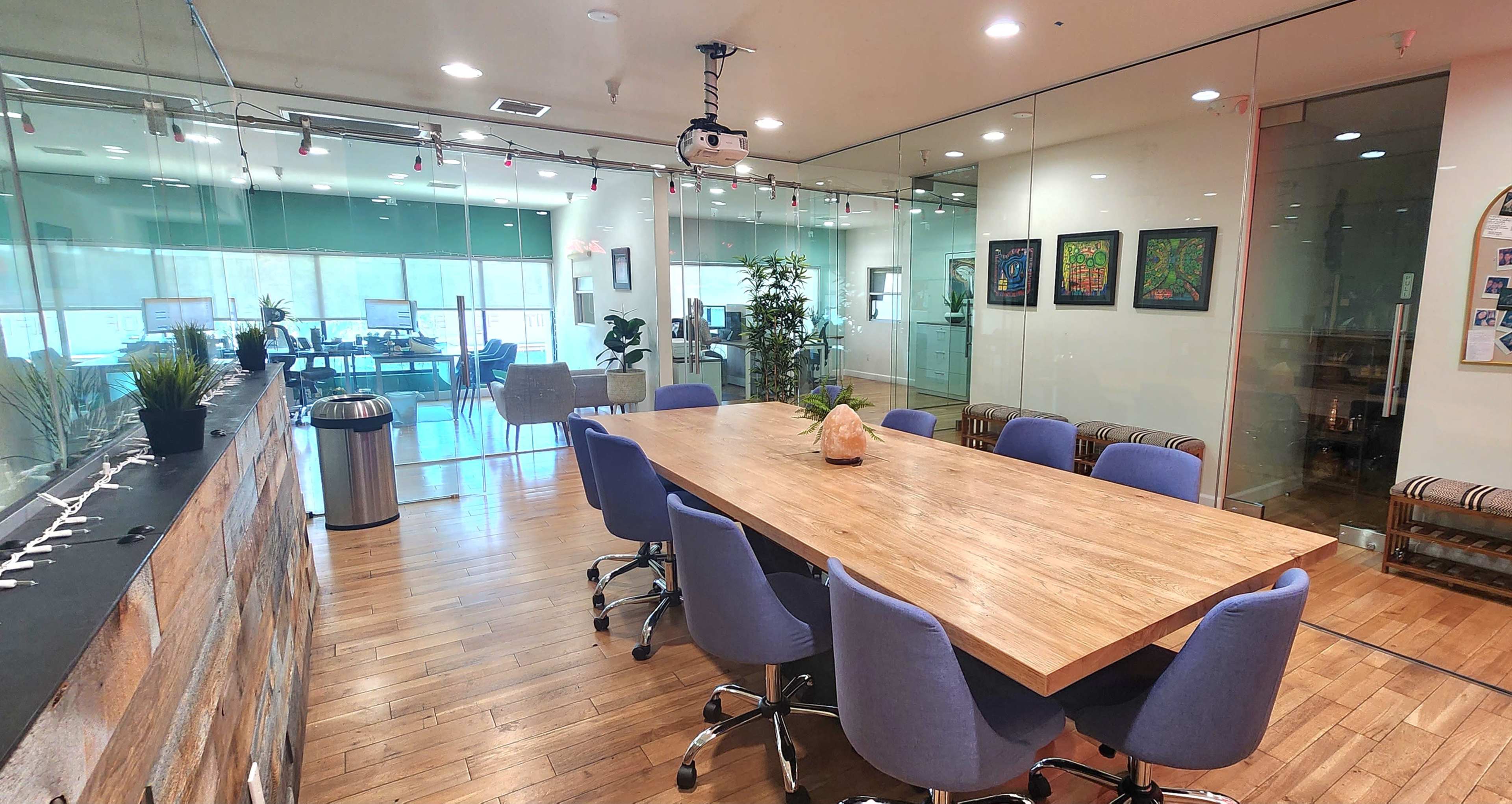 A modern conference room with a large wooden table surrounded by blue chairs, glass walls, and a projector mounted on the ceiling.