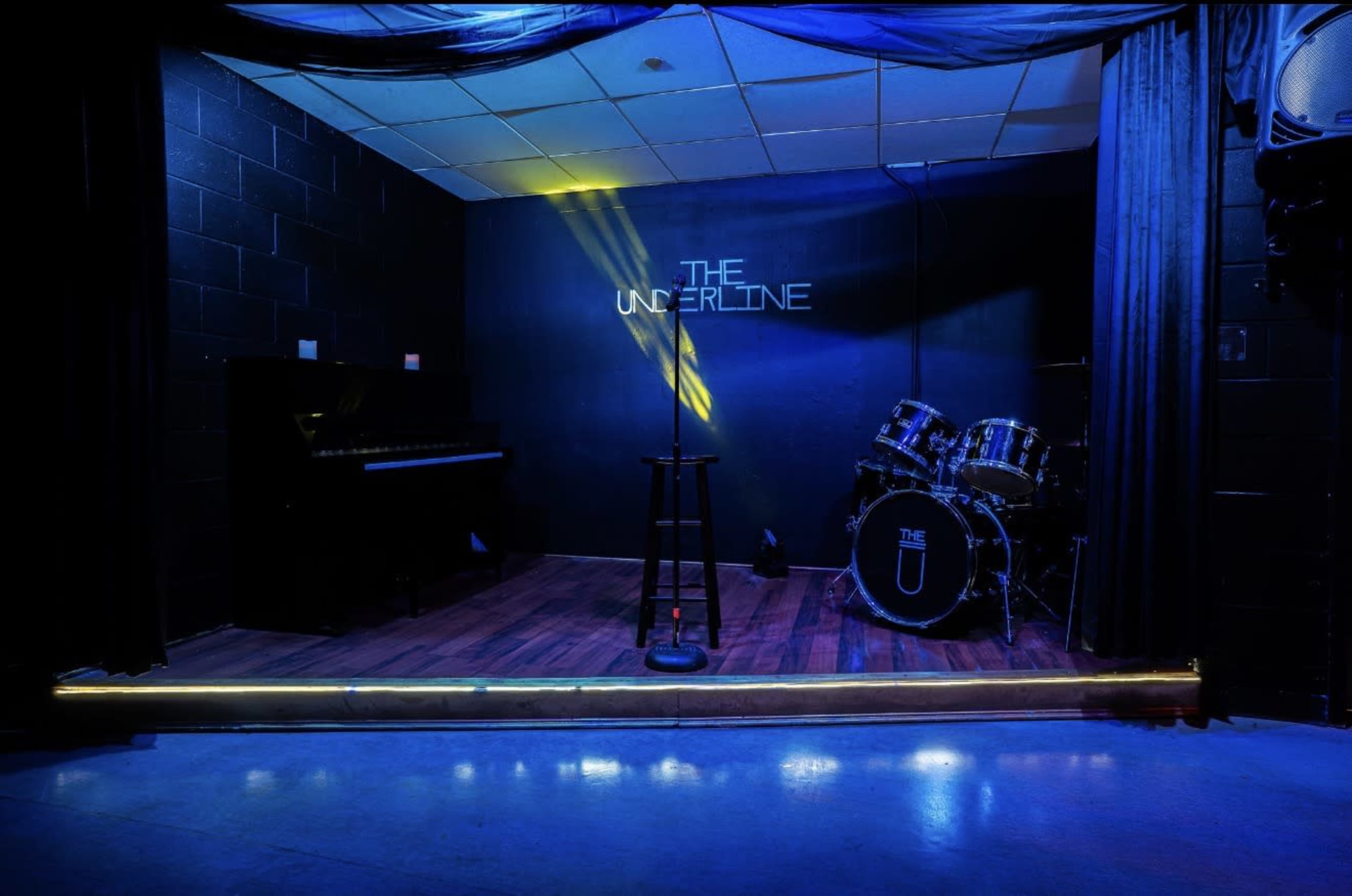 The image shows a performance stage with a microphone stand, a piano on one side, and a drum set on the other, all illuminated by blue lighting.