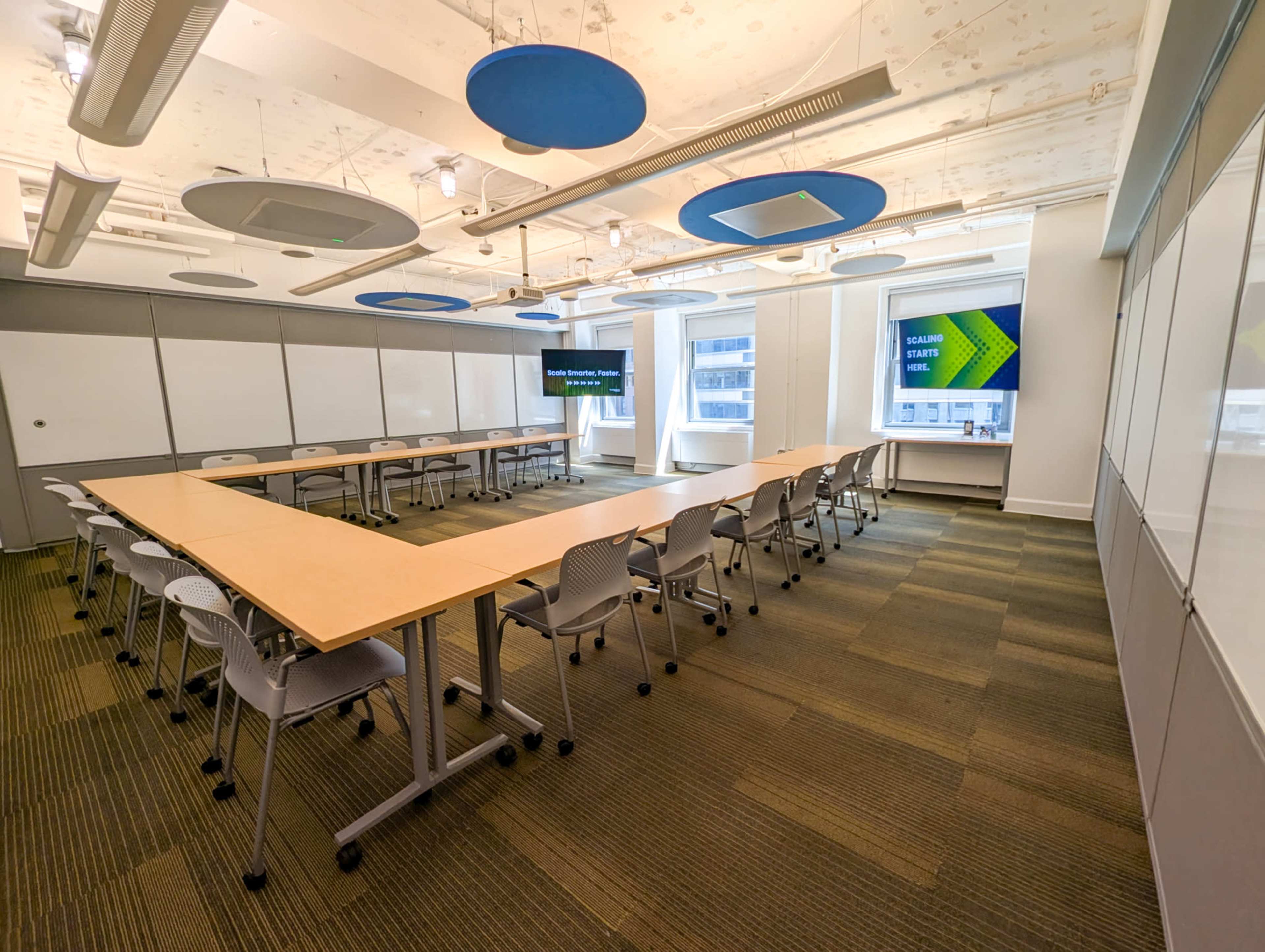 A modern conference room features a long table surrounded by chairs, with large screens on the walls and blue acoustic panels on the ceiling.