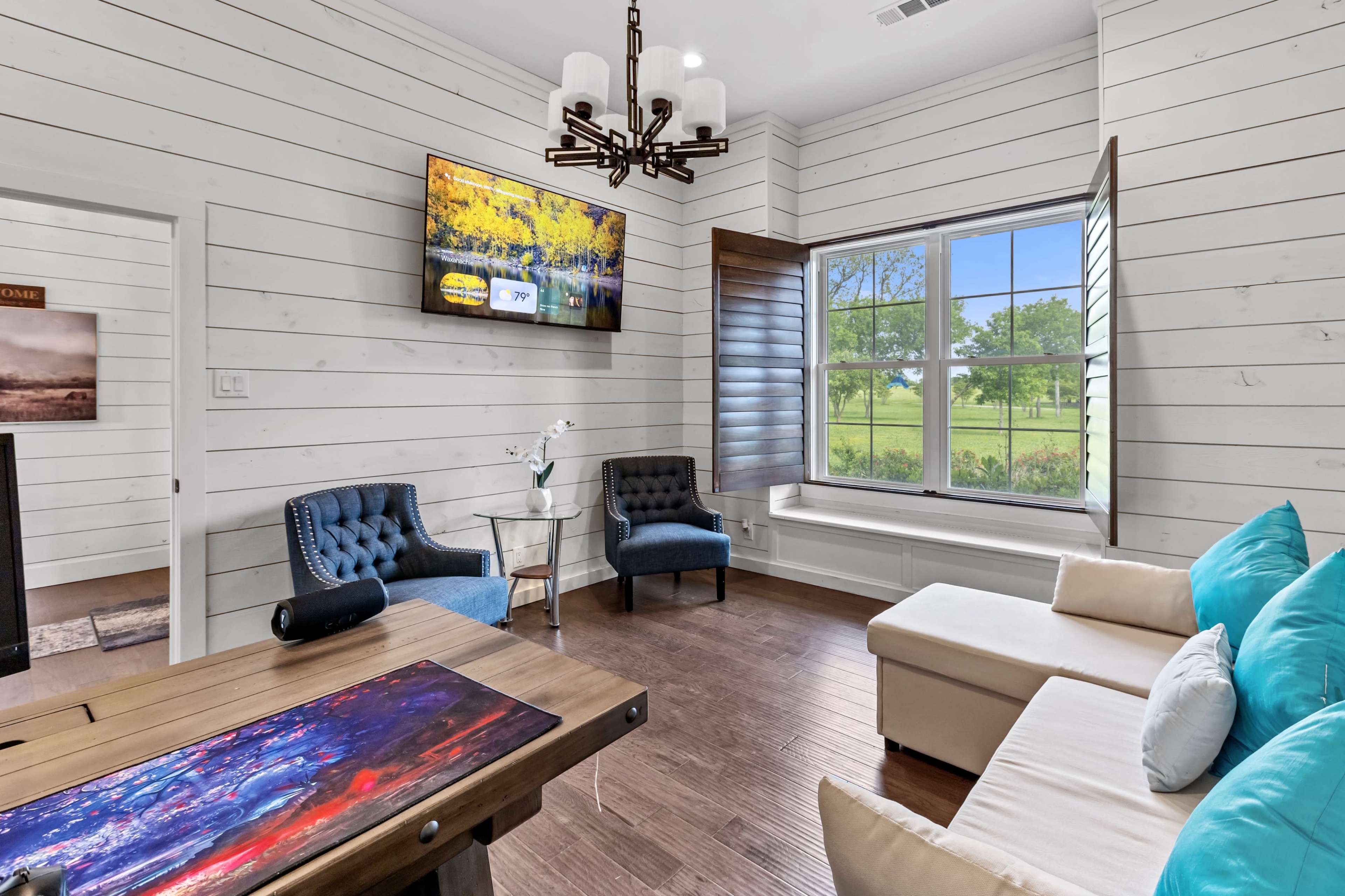 A cozy living room features a large window overlooking greenery, modern furniture, and a wall-mounted television.