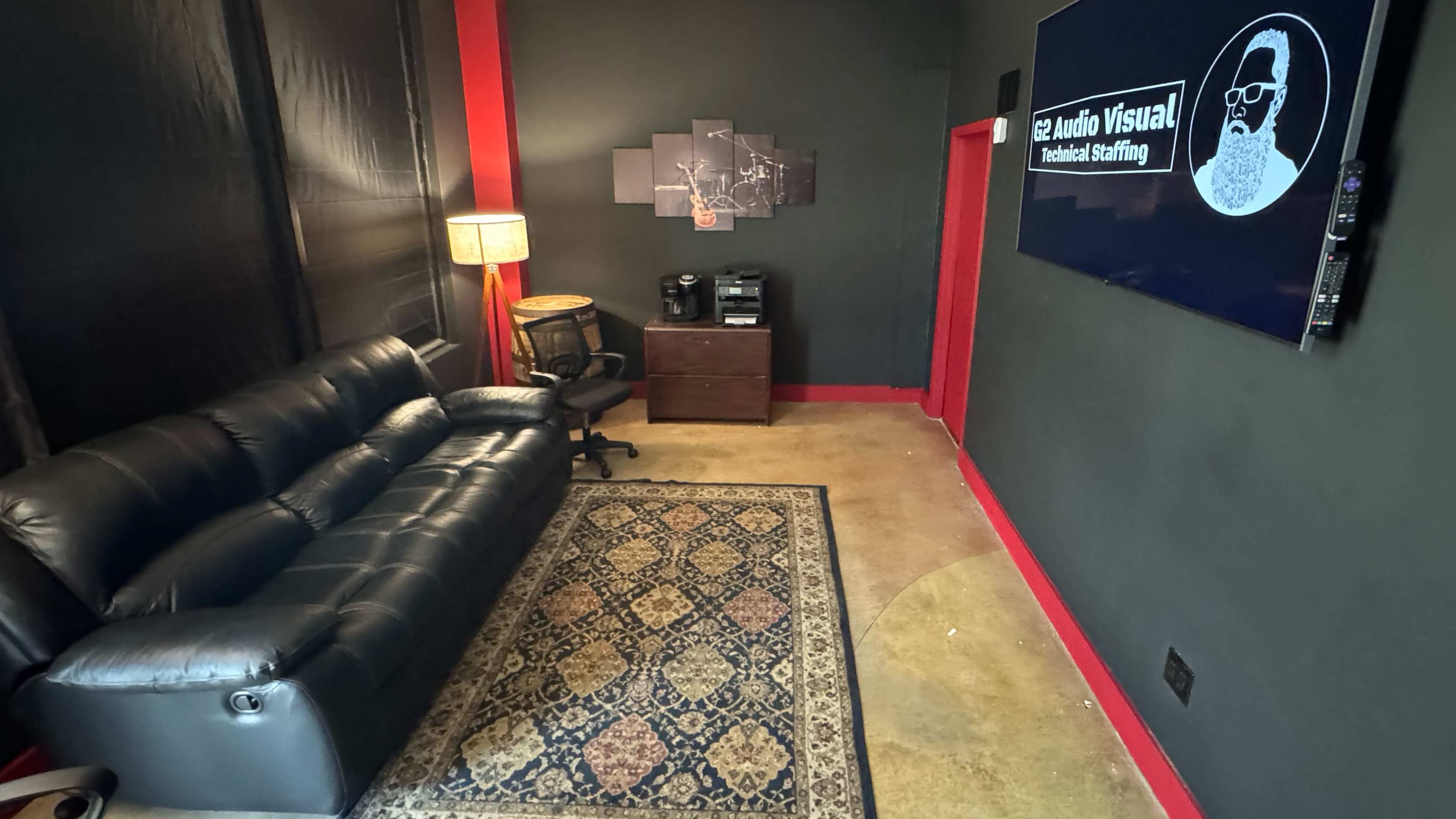 The image shows a small, modern room with a black wall, a leather couch, a rug, a lamp, a chair, and a mounted television displaying a logo.