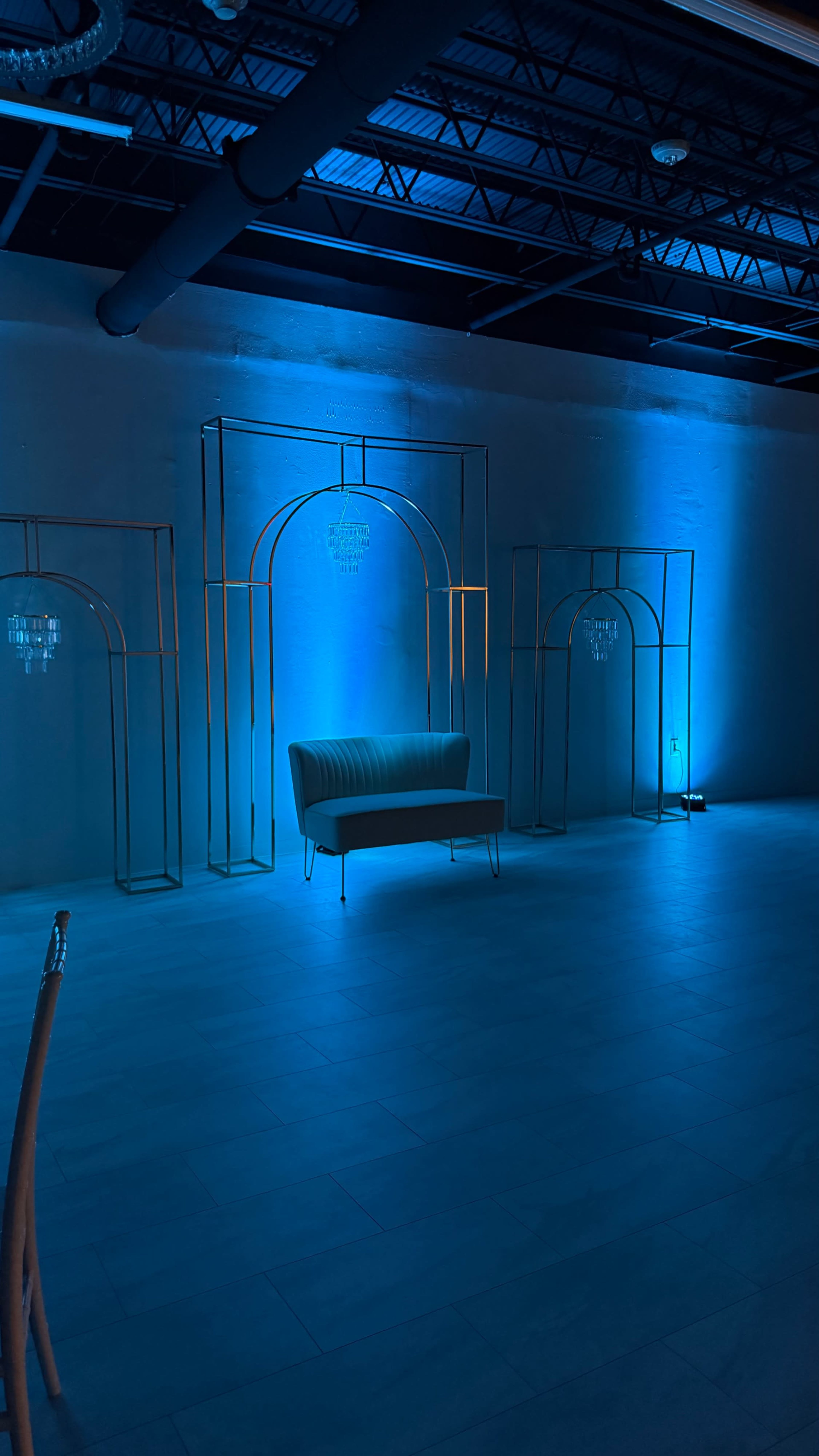 The image shows a dimly lit room featuring a green sofa positioned between three metallic arch structures, illuminated by blue lighting.