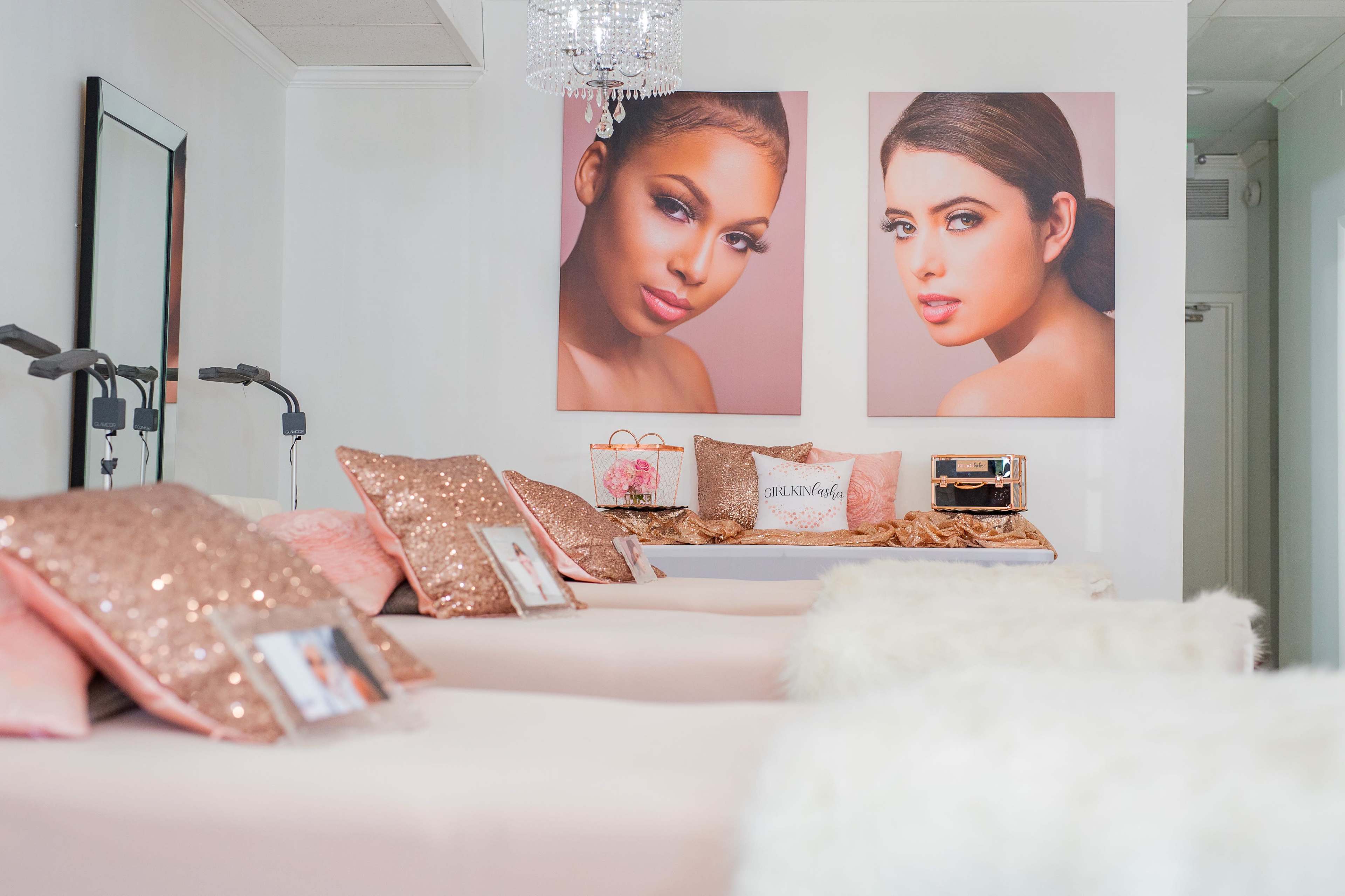 A beauty salon interior featuring two large portraits of women on the wall, with several treatment beds covered in white and pink decor, and sparkling decorative pillows arranged on them.